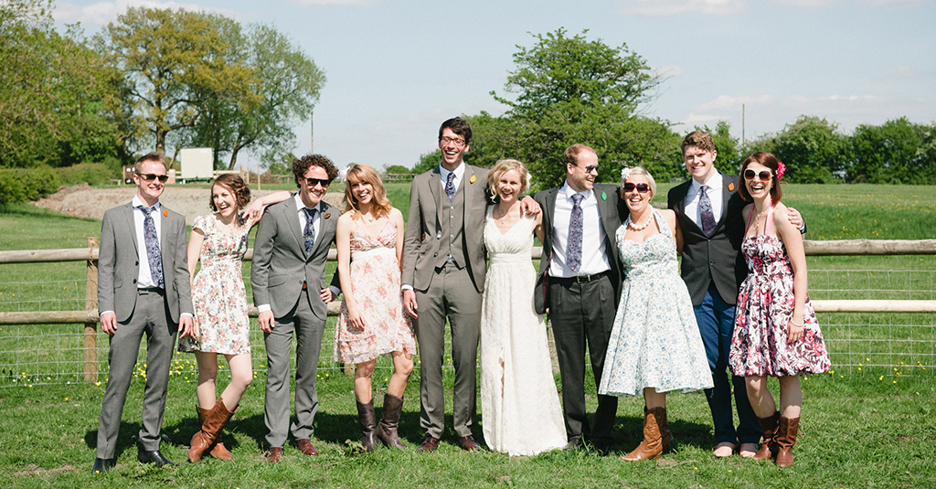 A bride and groom with eight friends by a fence on a grassy field. They're dressed casually for a modern wedding, with bridesmaids in pretty mismatched floral dresses. By Hannah Beatrice Photography