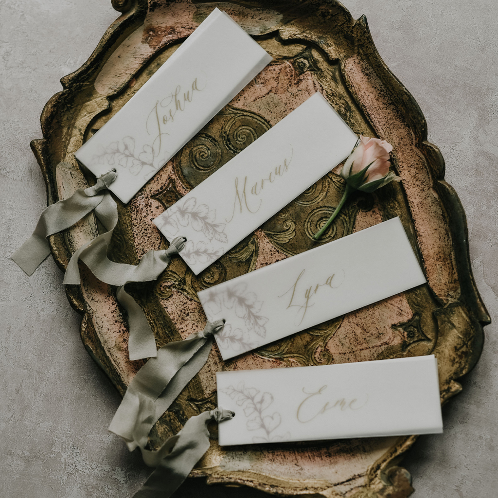 Four place name cards for a wedding arranged on an antique tray. They're written in modern calligraphy and have sage green ribbon ties. By Inkflower Press