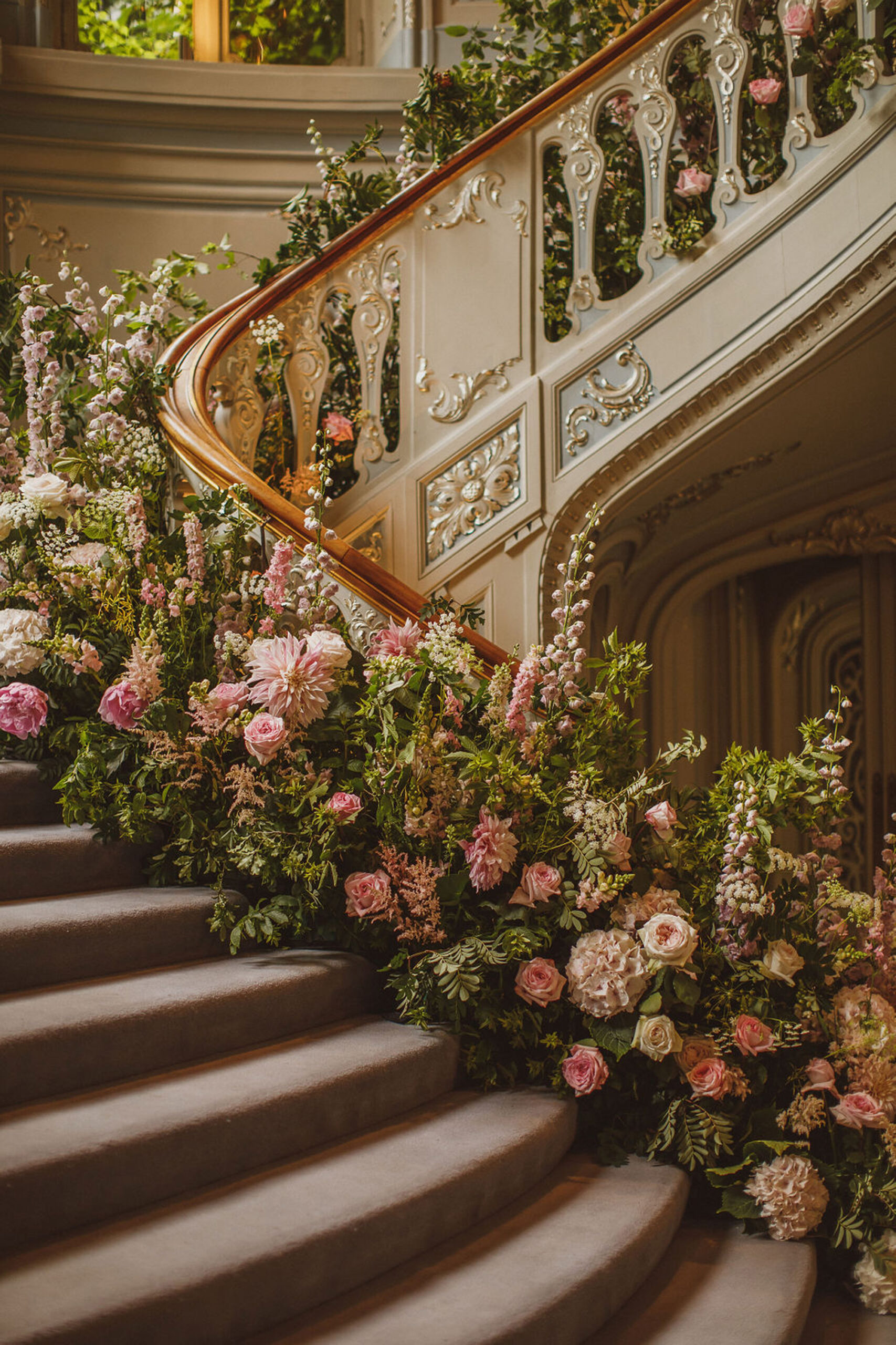 Luxurious pink and green florals almost cover the banister of a grand staircase at a wedding venue. By Niemierko