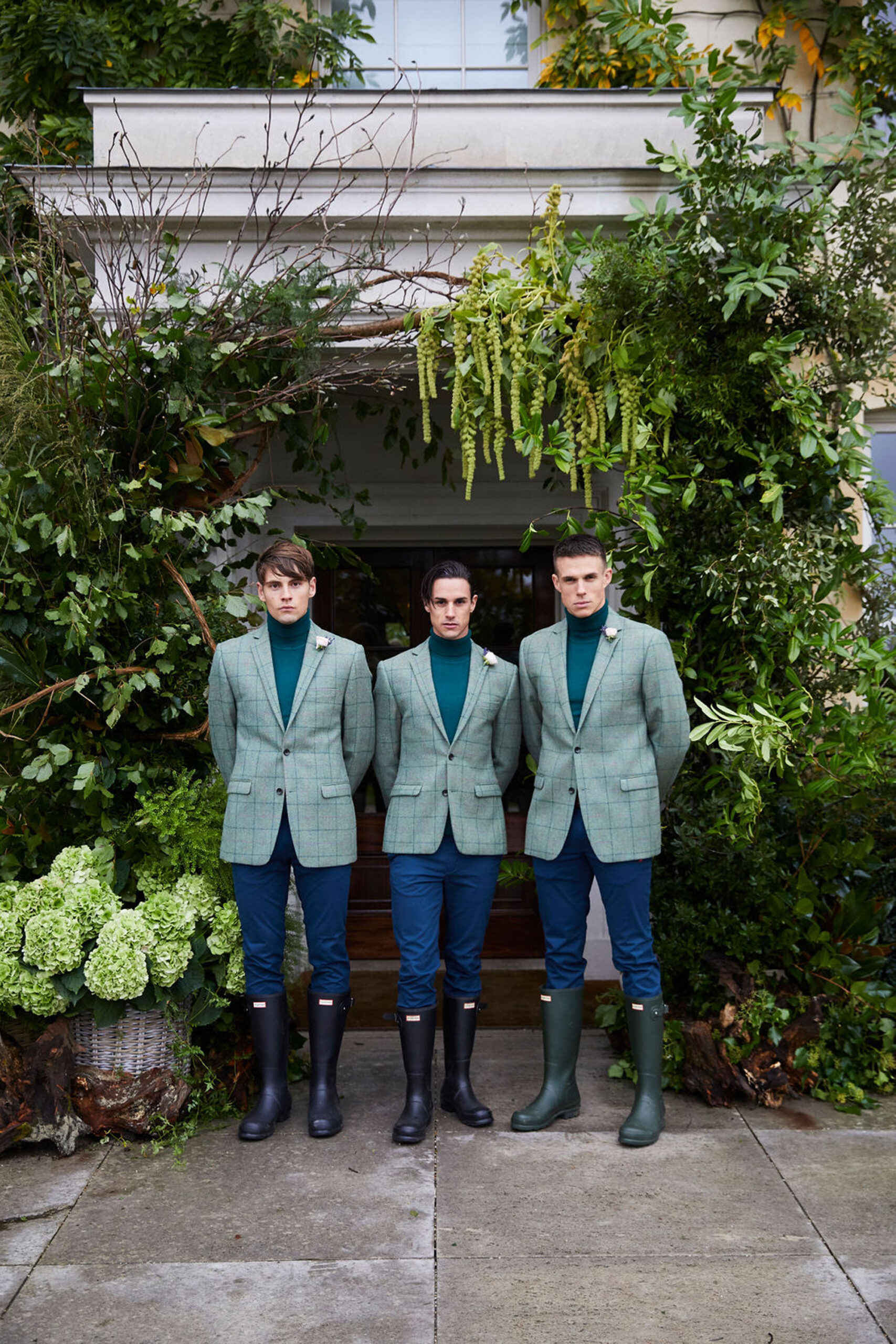 Three Niemierko hosts dressed in green jackets and boots stand amidst a display of greenery with leaves and luxurious green hydrangeas
