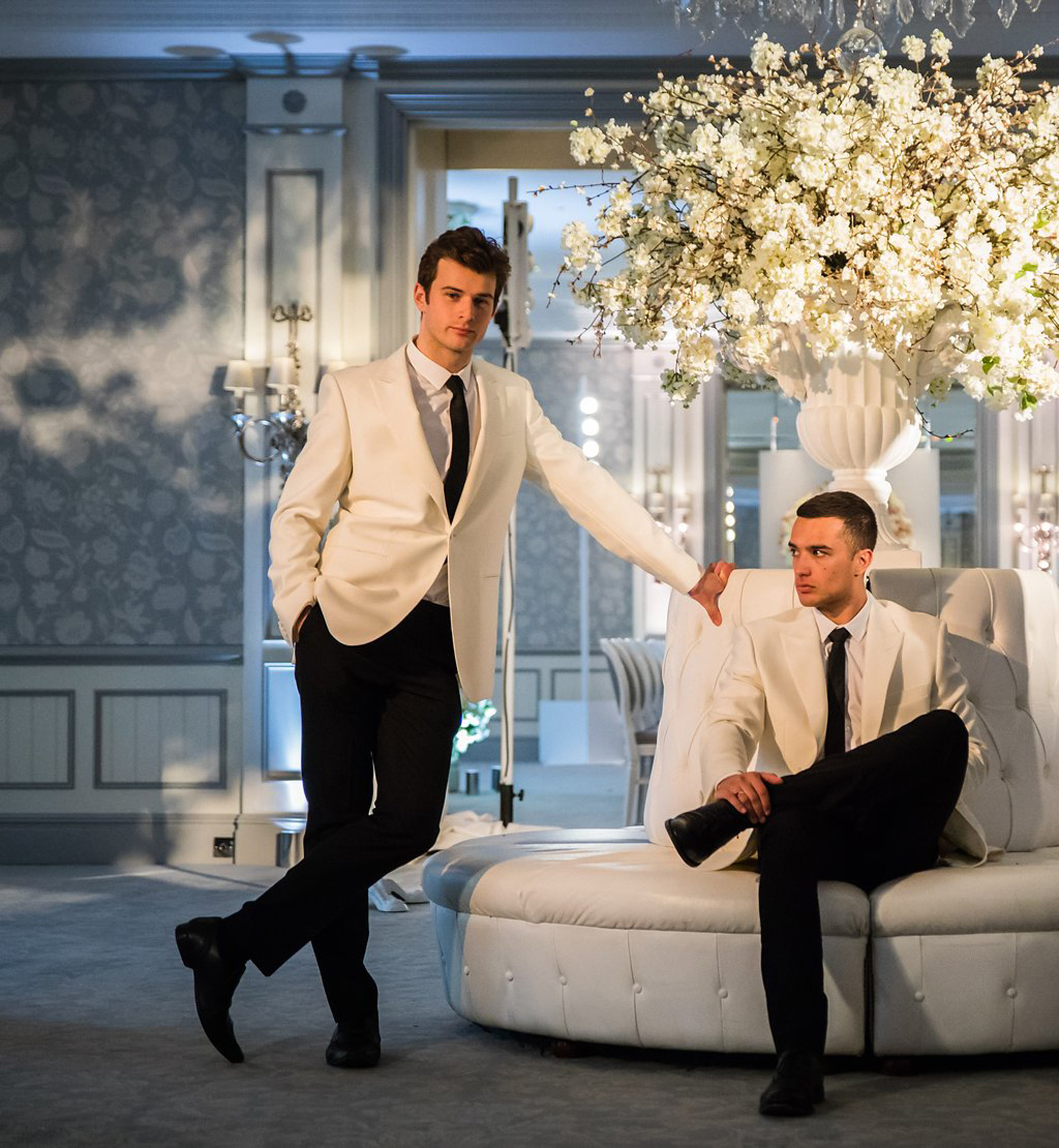 Niemierko hosts dressed in white suit jackets and black trousers pose for a photograph in a luxury hotel, seated on white leather below a luxurious pure white floral arrangement