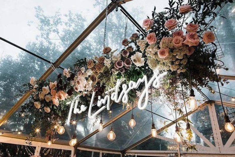 Neon wedding sign says The Lindsays hung from the frame of a glass roof and surrounded by florals and festoon lights