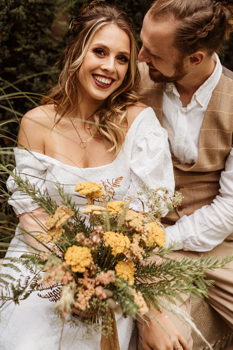 A couple pose for the camera. She's blonde, smiling and wearing an off-the-shoulder dress. He's looking at her. She holds a bouquet of wild yellow flowers. Photographer credit Sarah Hoyle, styling by Amethyst Weddings