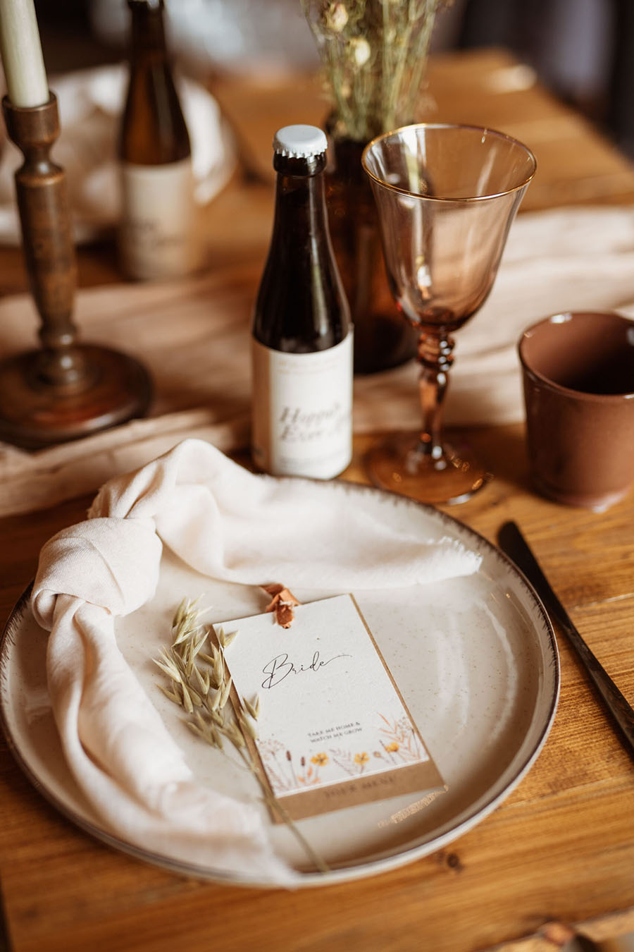 A wedding place setting on a wooden table. A plate with a knotted napkin and place name card, with a brown glass bottle and smoky brown wine glass. Photographer credit Sarah Hoyle, styling by Amethyst Weddings