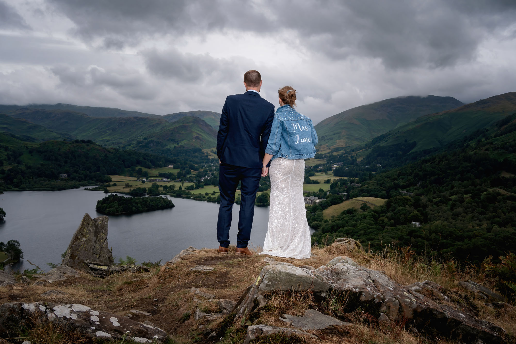 A couple stand on a mountainside looking over a lake. She's wearing a long white skirt and denim jacket. He's in a dark suit. By Jaye-Peg Photography in Cumbria