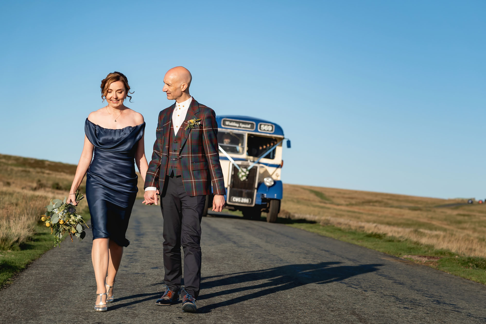 A couple walk along a country lane with a vintage bus parked behind them. She's wearing a navy blue satin dress and he's in a dark suit. By Jaye-Peg Photography in Cumbria