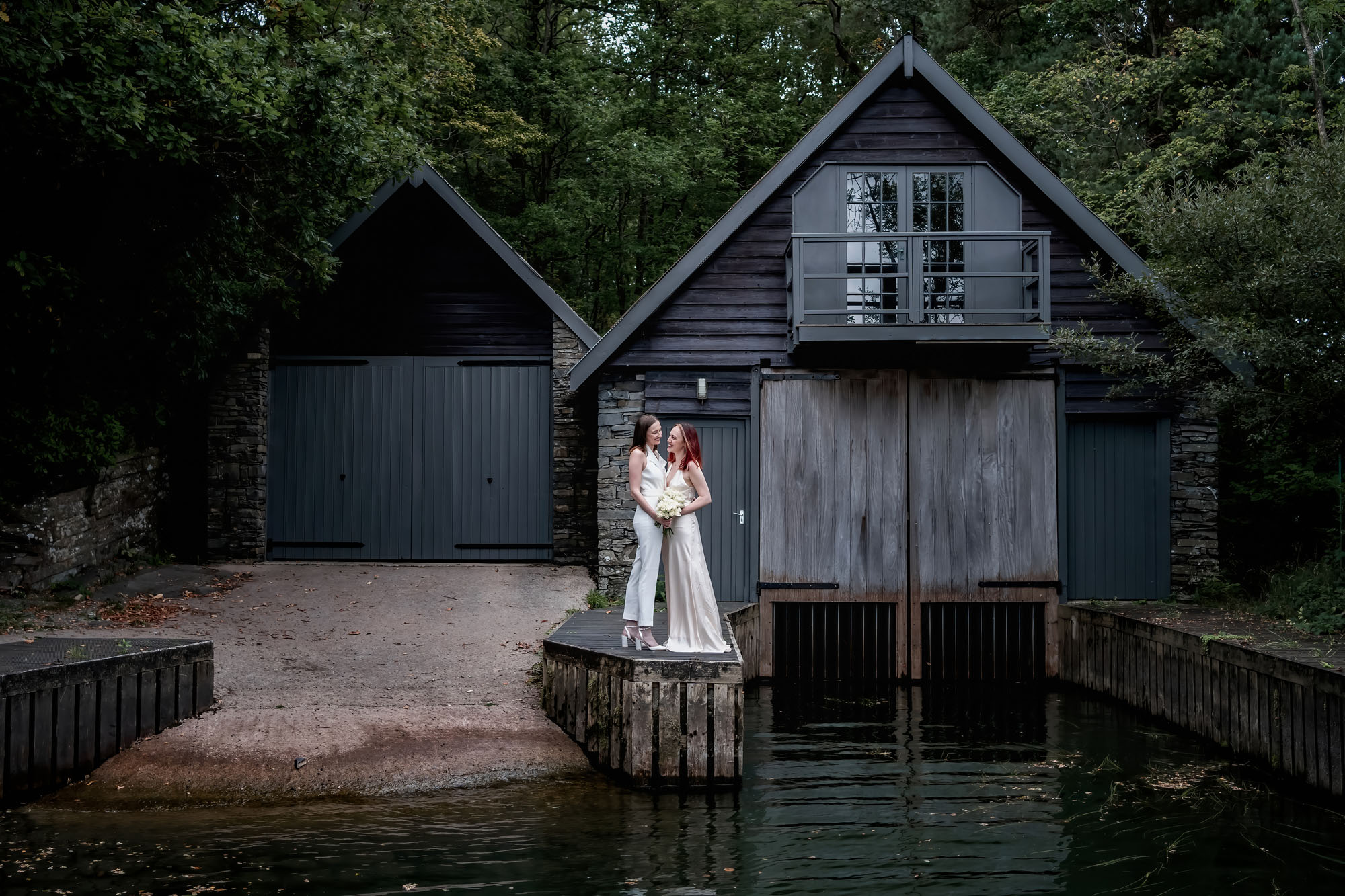 A couple stand at the end of a jetty by a boathouse. They're wearing white dresses and hugging. By Jaye-Peg Photography in the Lake District