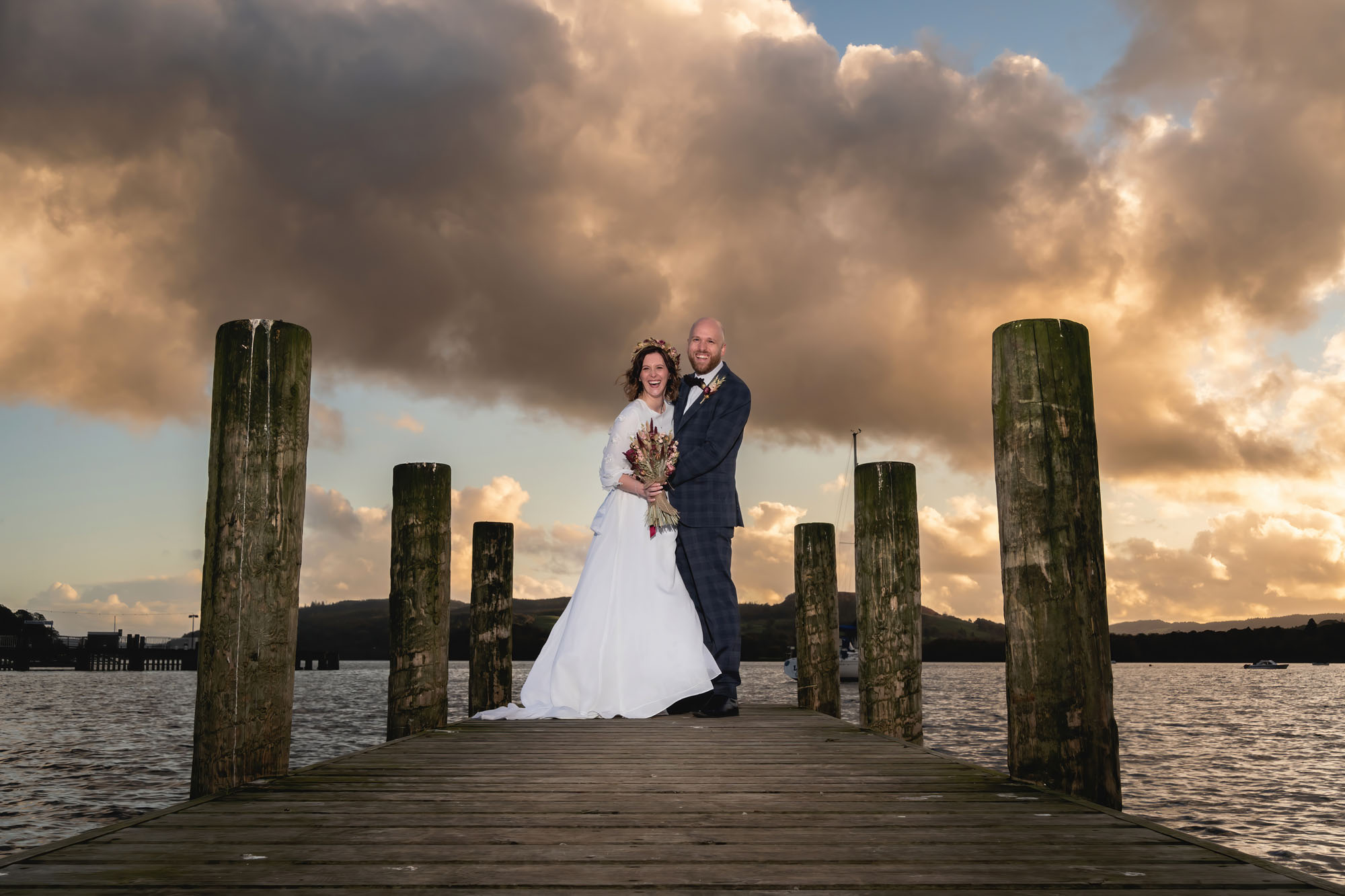 A couple stand on a jetty at sunset. She's wearing a white wedding dress and holding a bouquet. He's in a dark suit.