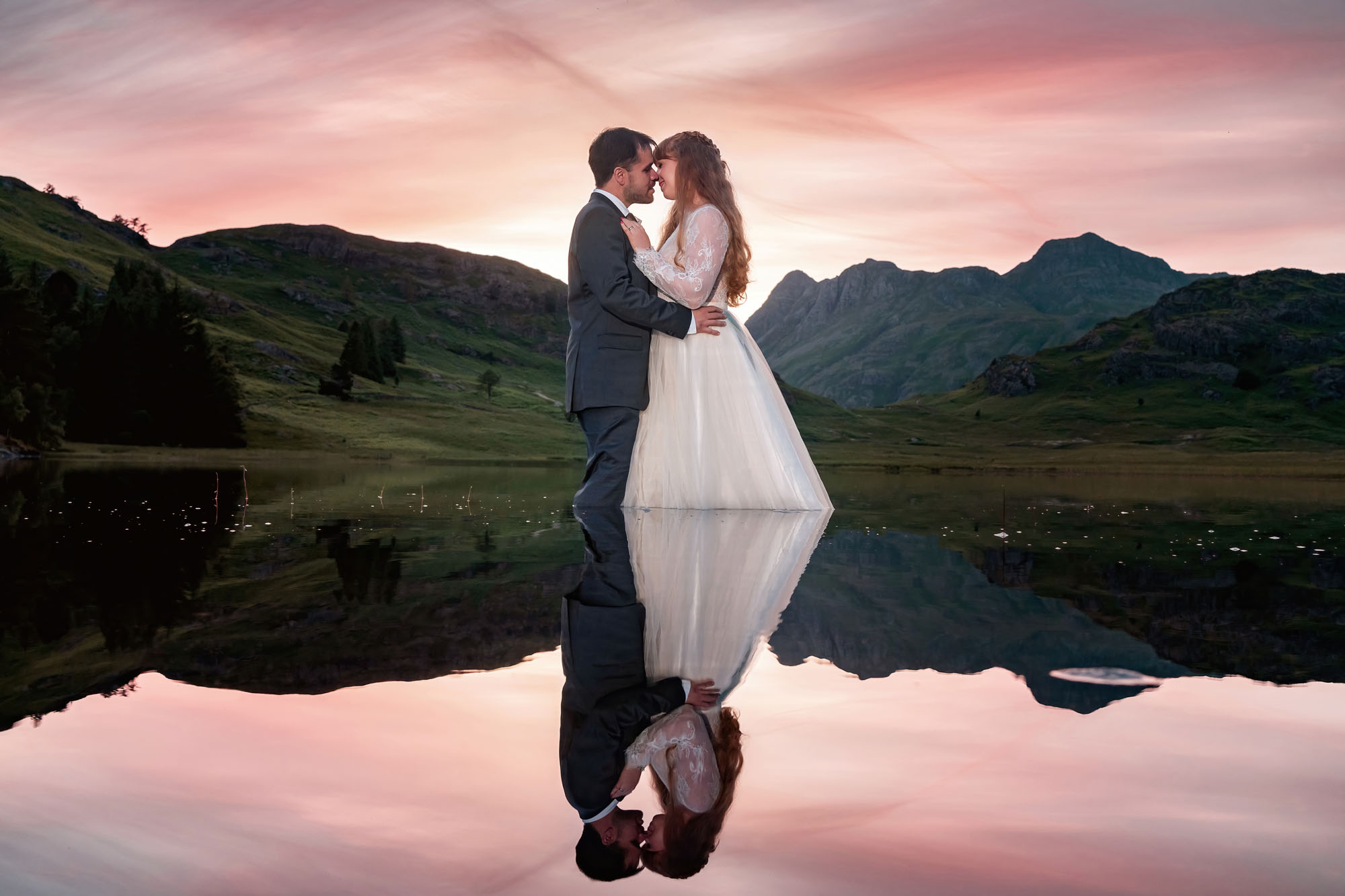 A couple share a kiss at sunset and their reflection is in the Lake before them. Beautiful Lake District wedding photography by Jaye-Peg Photography