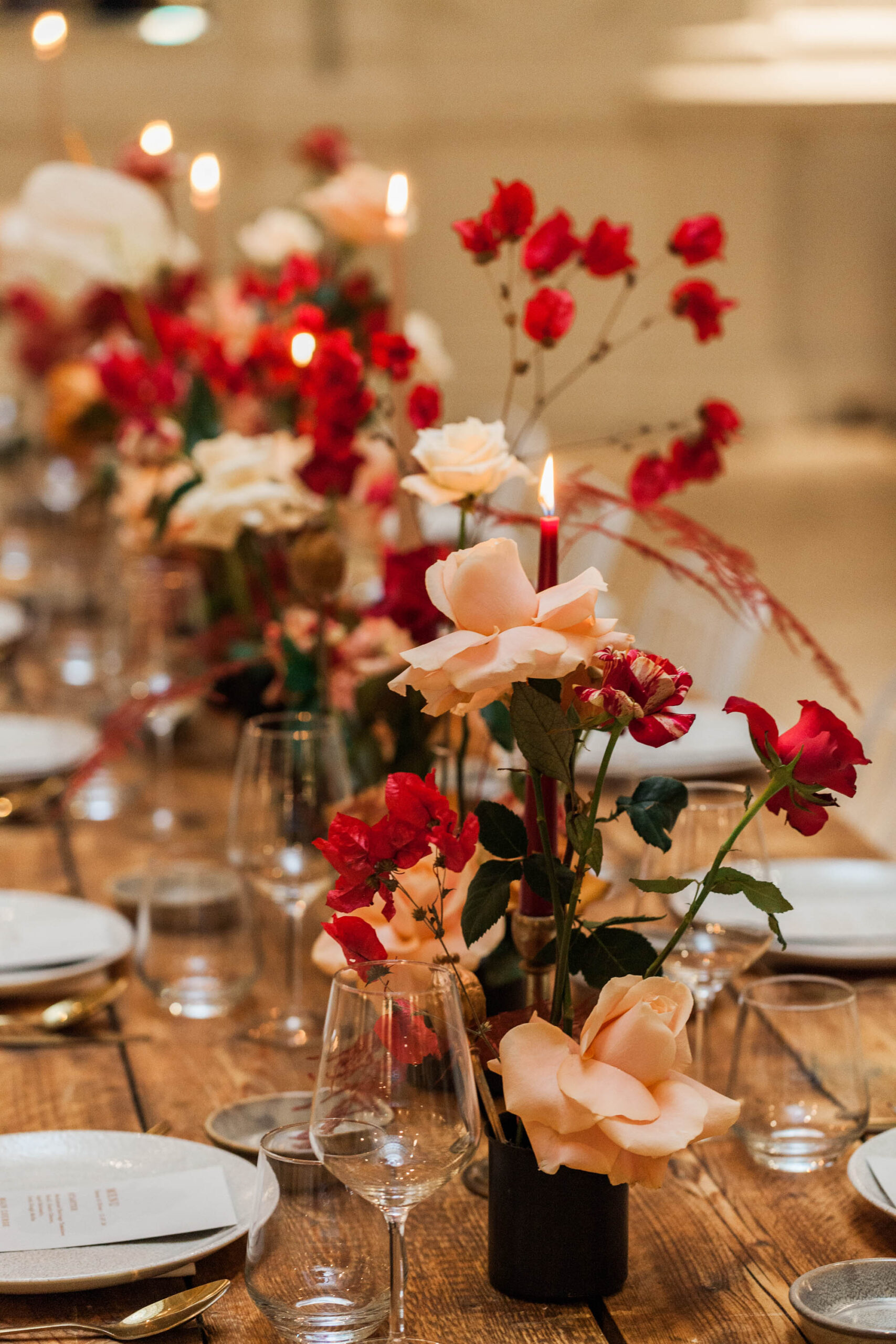 A wedding table laid with white crockery and delicate glassware, with candles and flowers in reds, corals and cream