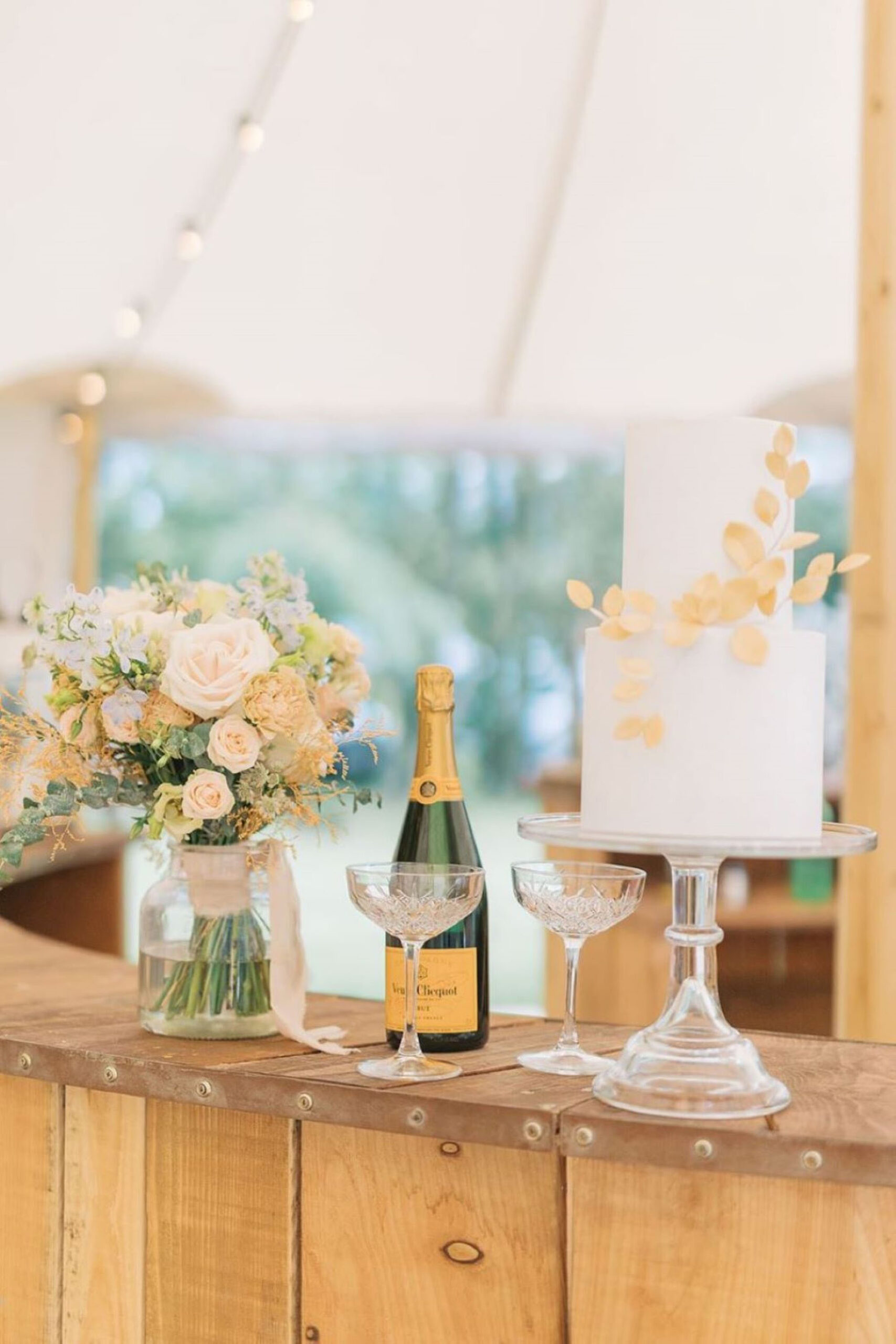A wooden bar in a wedding Sperry tent, with a pale coral bouquet in a vase, a bottle of Veuve Cliquot champagne and two glasses, and a simple two tier wedding cake with gold leaf decoration