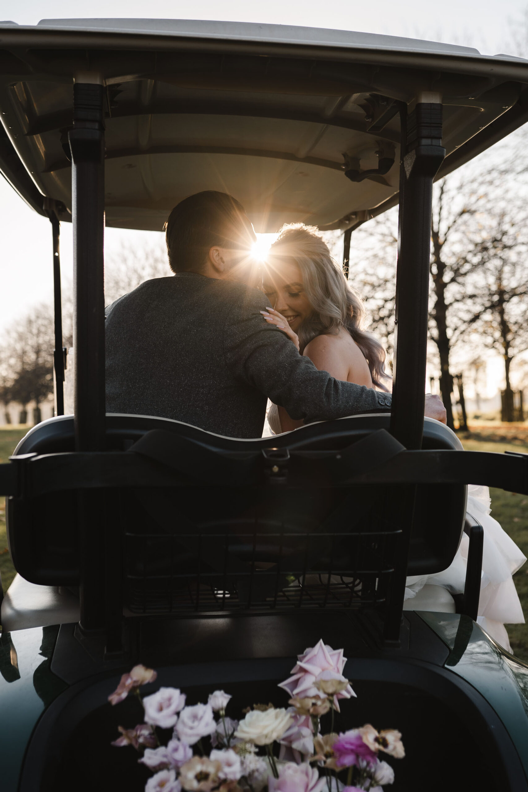 Wedding couple in a golf cart. She's leaning into his shoulder for a forehead kiss, and the sunlight is shining between them. By Karolina Photography