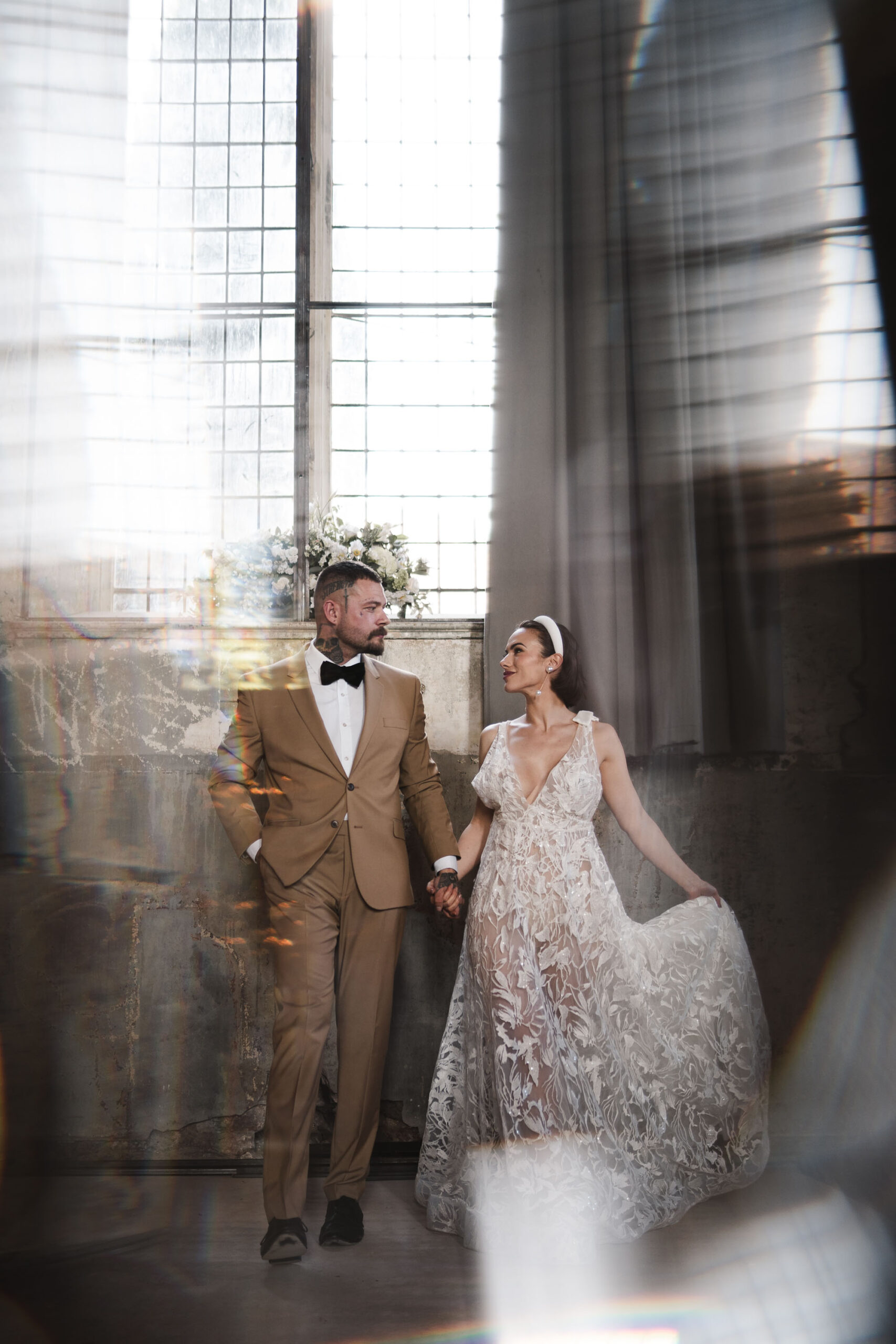 Wedding portrait from By Karolina. A bride and groom stand before a window. He's wearing a tan suit and black bowtie. She's in a patterned white dress which she's holding out to the side.