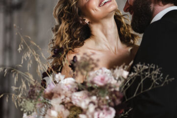 Photograph of a bride and groom. She's carrying a bouquet of pink flowers and grasses, and has lifted her head so he can kiss her neck. By Karolina Photography