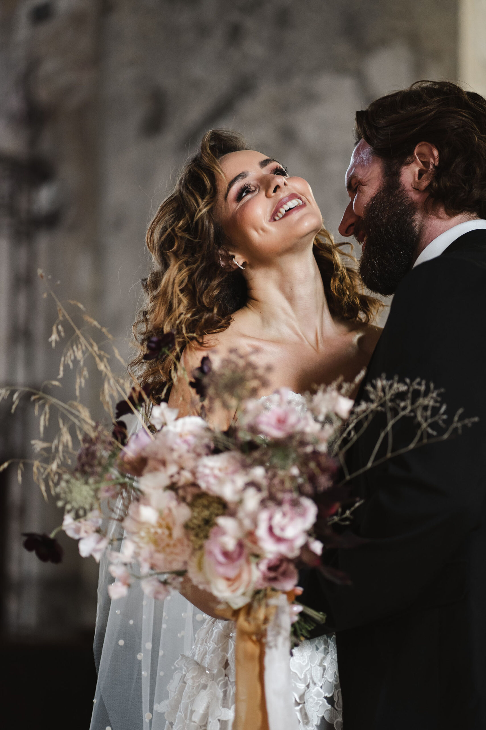 Photograph of a bride and groom. She's carrying a bouquet of pink flowers and grasses, and has lifted her head so he can kiss her neck. By Karolina Photography