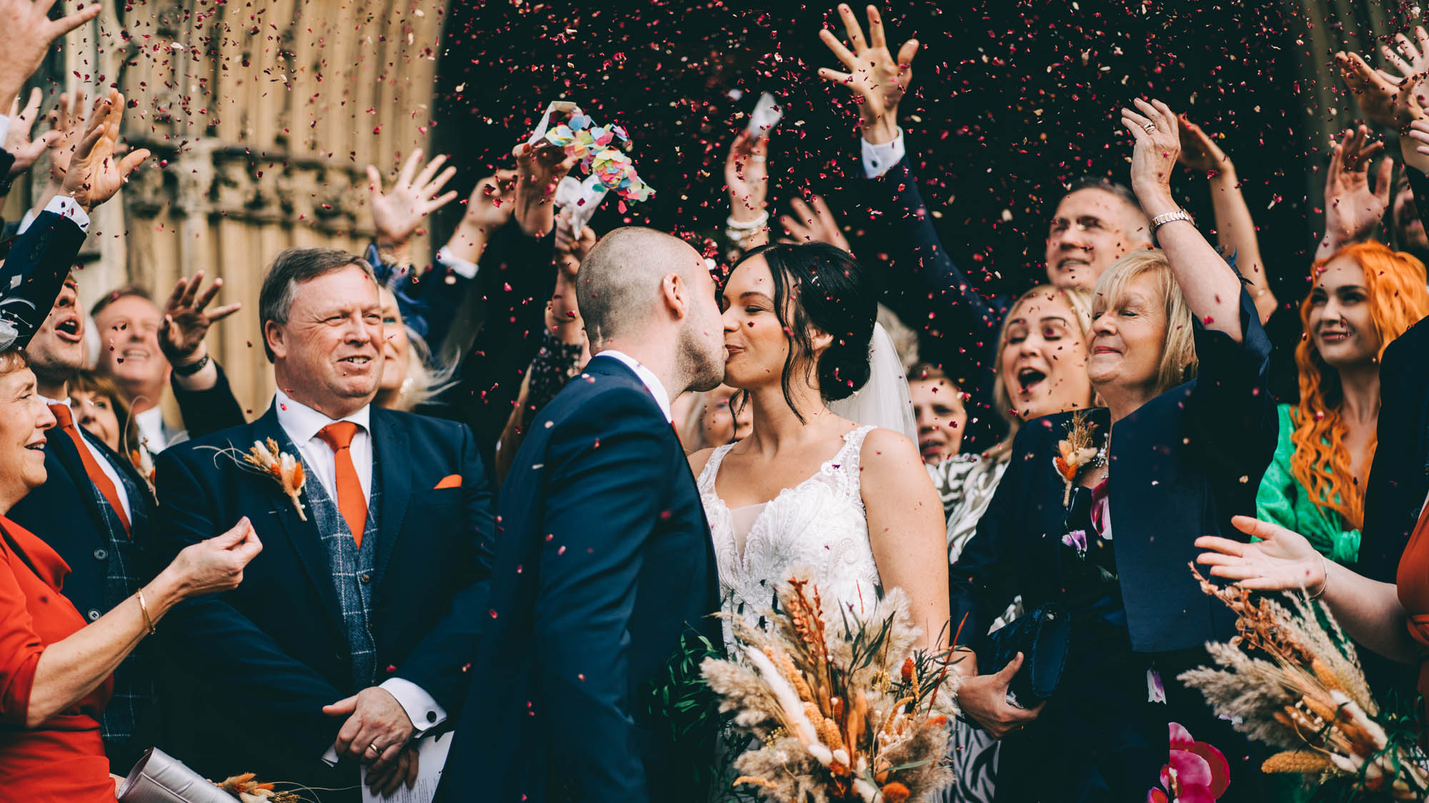 A groom and bride kiss as guests throw confetti. By Abraham Photography
