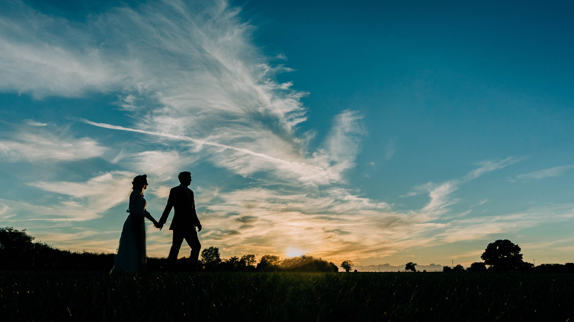 A couple walk along the horizon in the countryside as the sun sets. The sky is blue with vapour trails. By Abraham Photography
