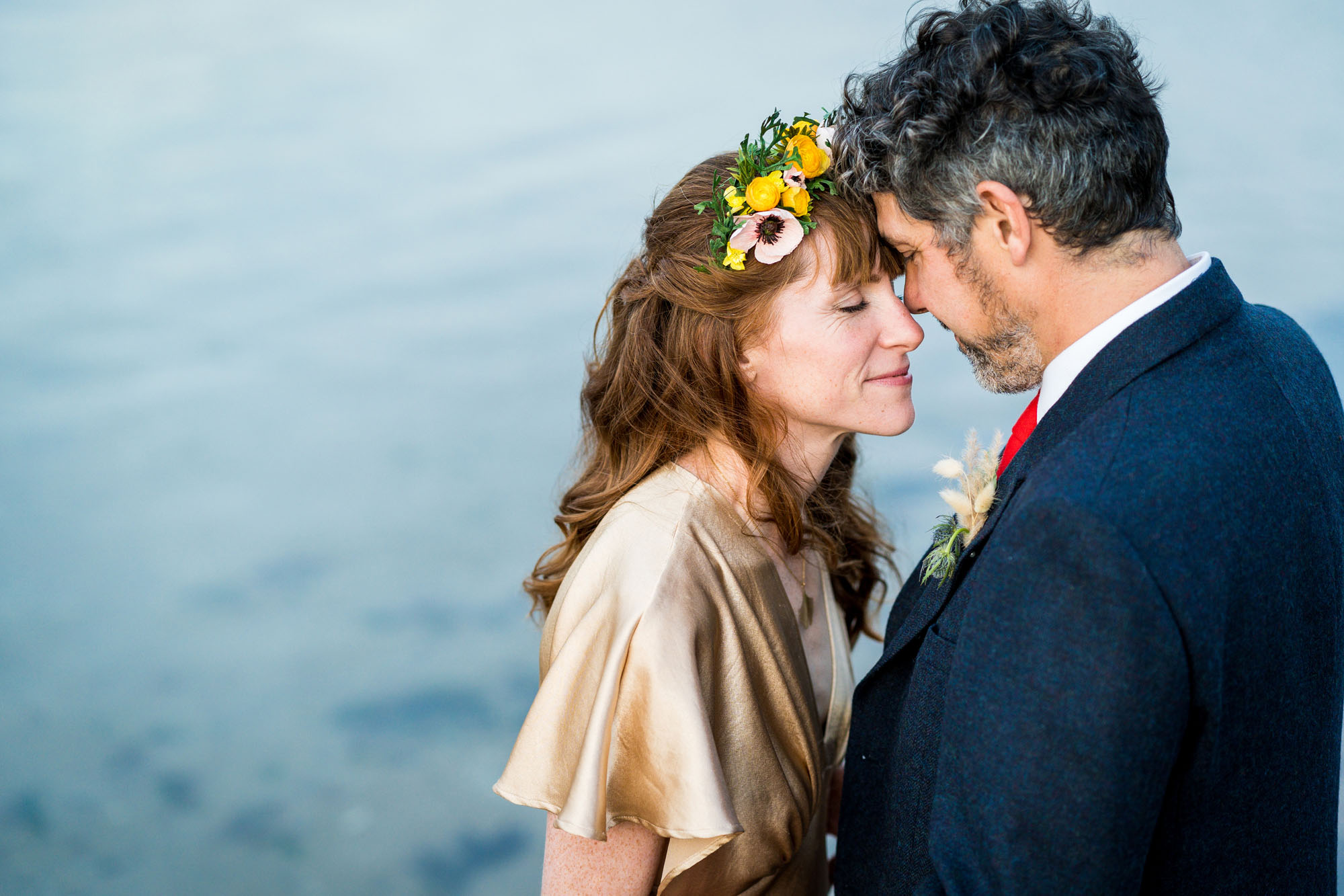 Lisa and Duncan planned an alternative wedding to suit them. Lisa's wearing a gold dress and a paper flower hair accessory, and Duncan's in a blue jacket with red tie. They're touching foreheads and smiling, with the sea behind them. By Benjamin Toms Photography in Kent
