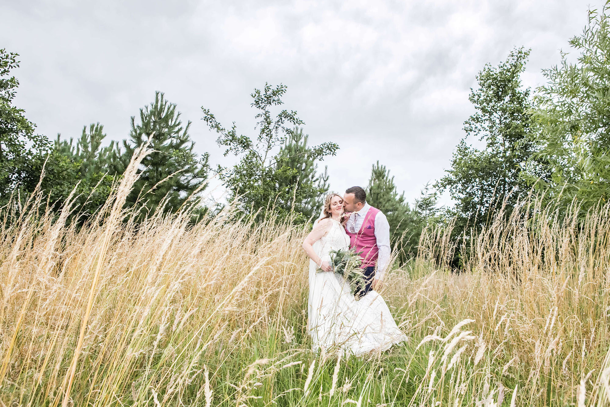 A bride and groom on their wedding day, in a field of long grass. The camera angle is low and there are green trees behind them. By Erika Tanith Photography