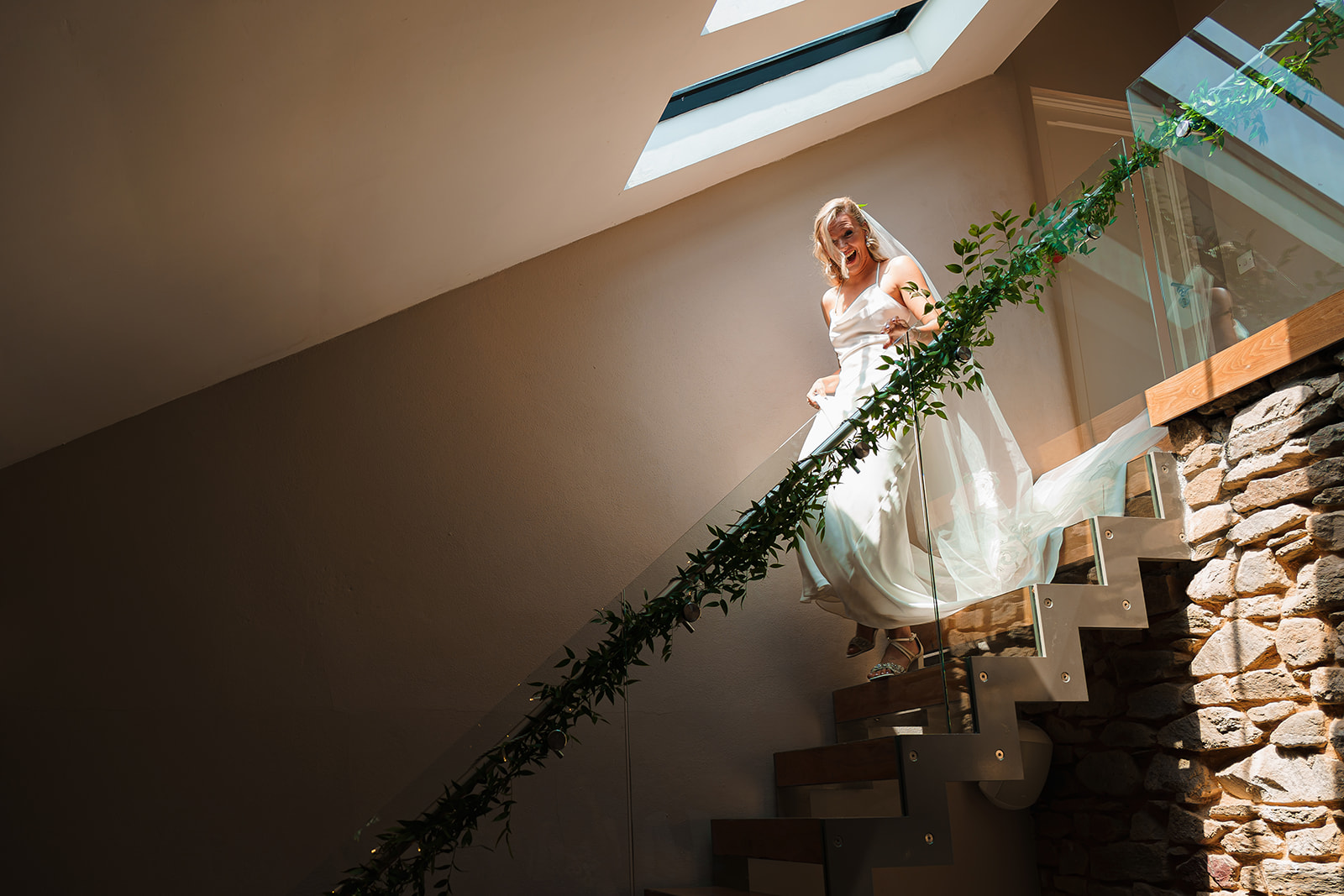 A bride walks down the stairs under a skylight which pours beautiful soft light onto her. The banister is styled with foliage. By Tracey Warbey Photography