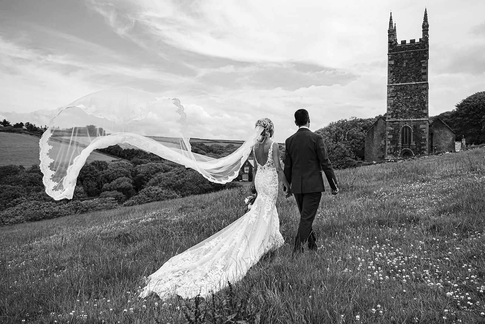Black and white photo of a couple walking across a field to a church in the distance. They're in wedding outfits and her veil is blowing in the wind. By Tracey Warbey Photography