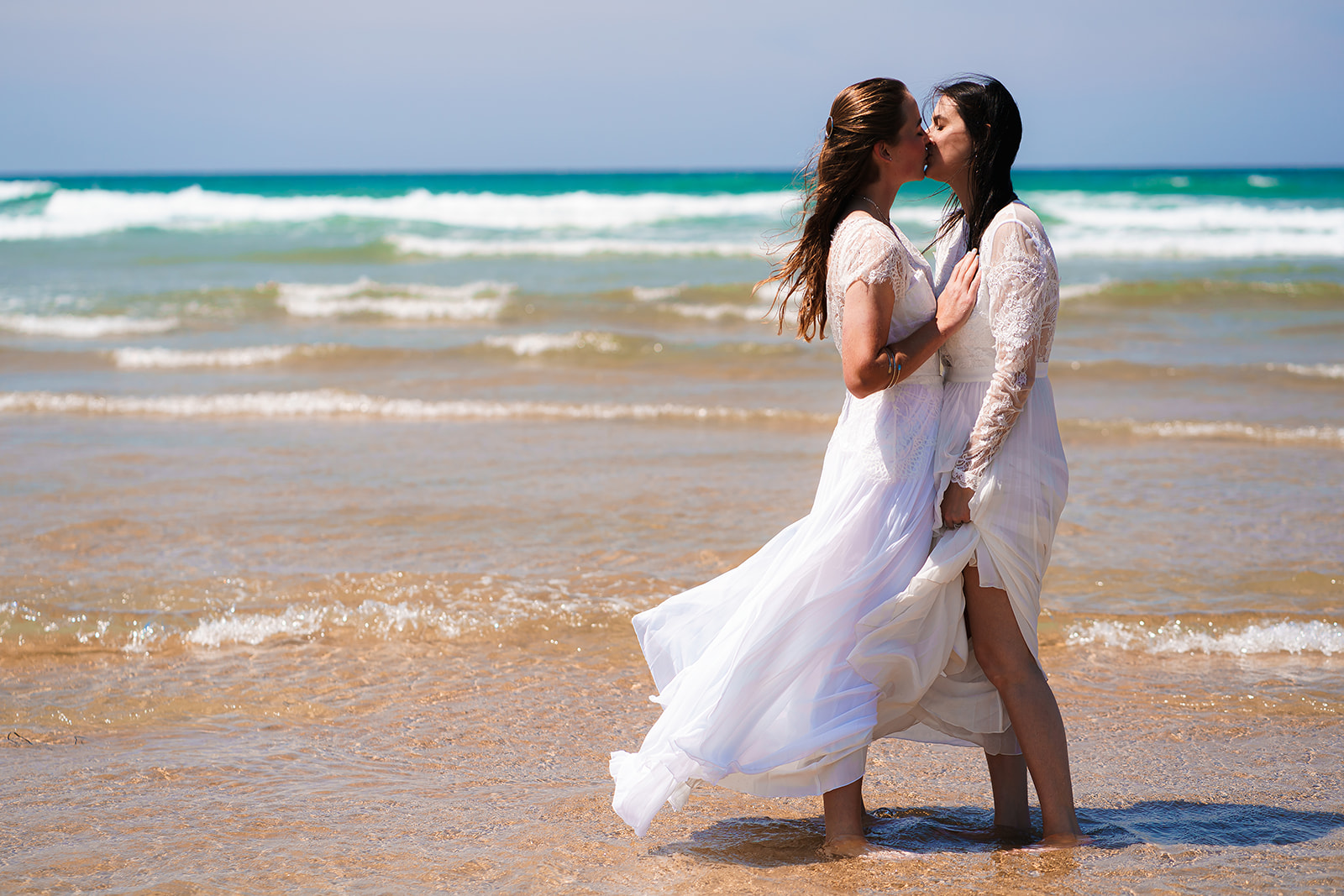 An LGBTQ couple kissing on a beach. They're both in wedding dresses and the waves are lapping onto their feet. By Tracey Warbey Photography