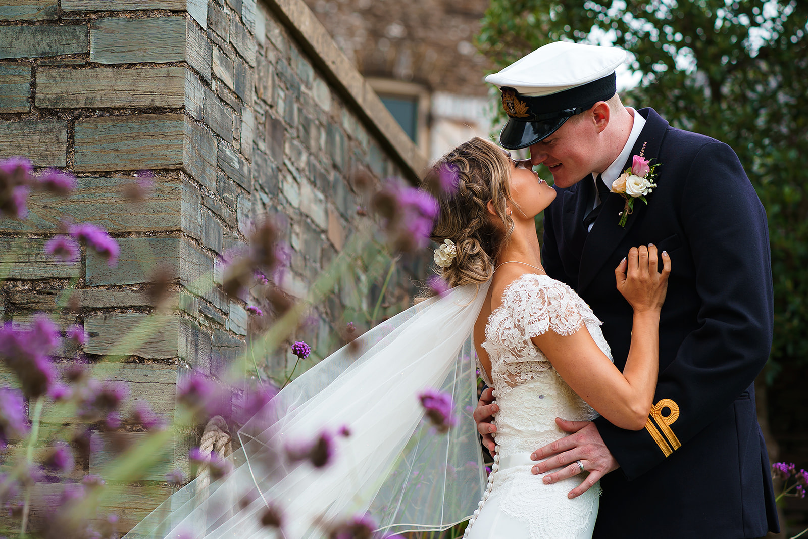 A couple are kissing on their wedding day. She's wearing a short sleeved white dress with lace and a veil. He's in a military uniform. By Tracey Warbey Photography
