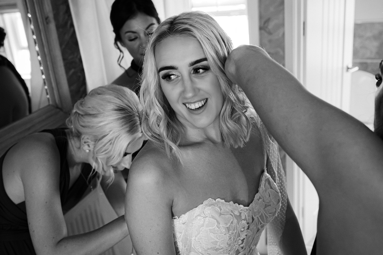 A bride getting ready for her wedding, with bridesmaids helping fasten the back of her wedding dress. A black and white photo by Tracey Warbey Photography