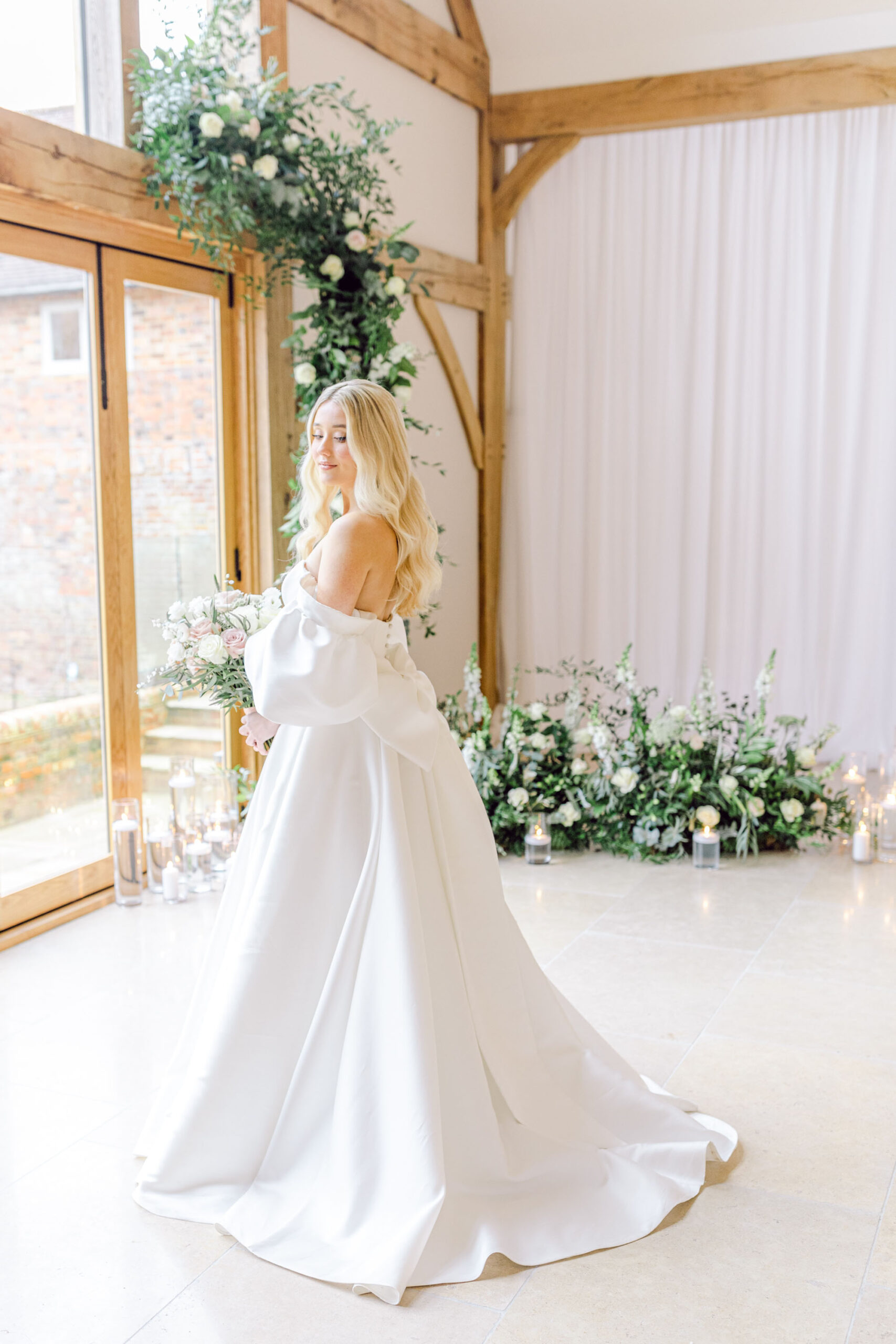 A bride model poses in a full skirted off the shoulder dress. There's a floor to ceiling window in front of her, surrounded by foliage. Taken at luxury wedding venue Greentrees Estate by Natalie D Photography