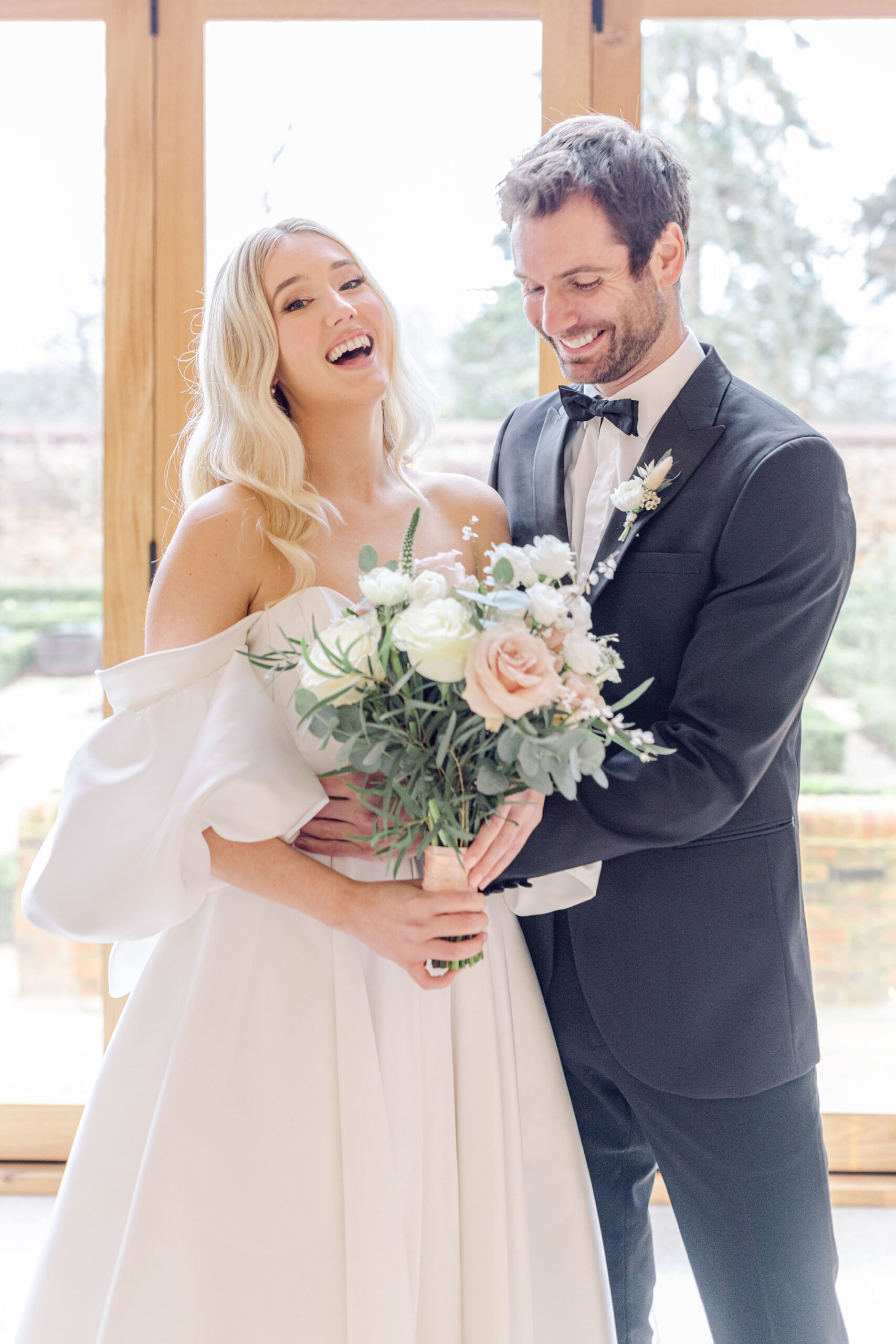 Model couple laughing together on a luxury wedding photoshoot. She's wearing a white dress and he's in black tie. They're holding a bouquet of white and blush roses. Image by Natalie D Photography