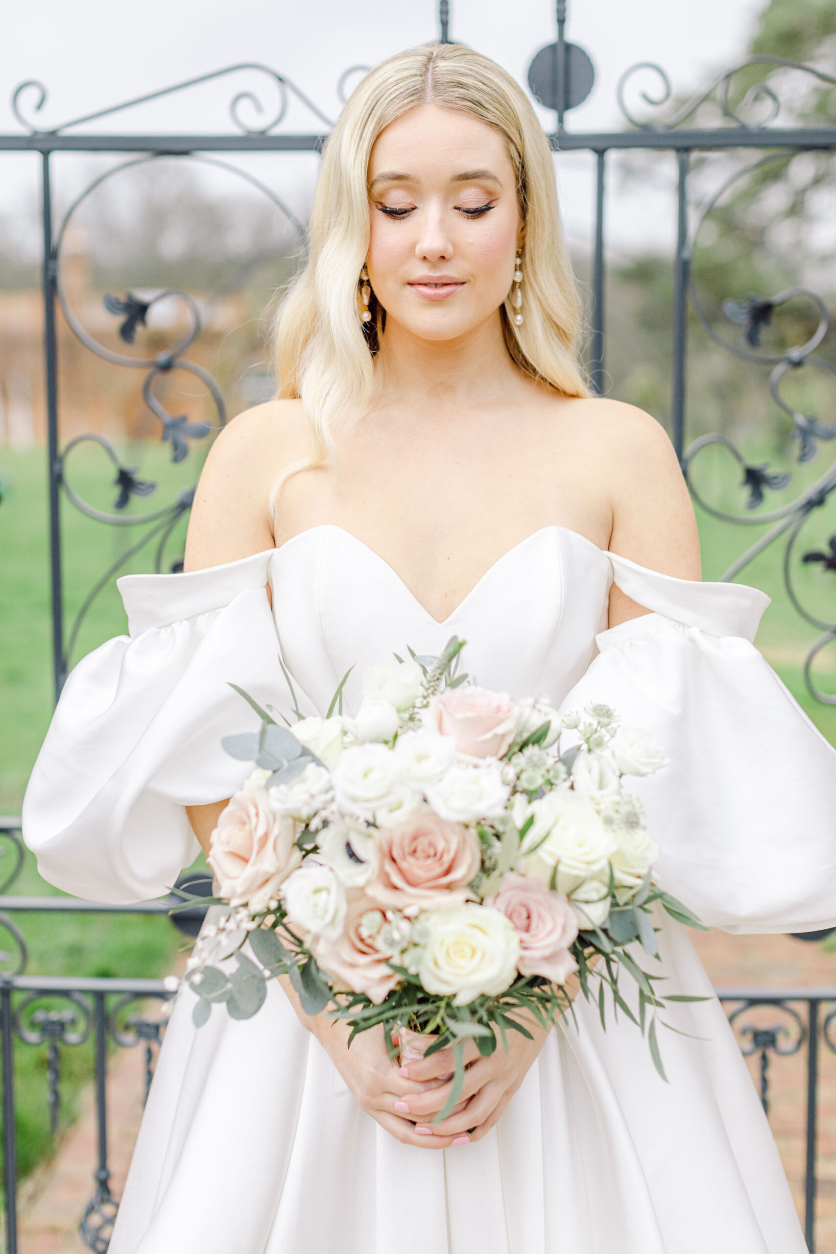 A model bride poses with a bouquet of white and blush roses. She wears a luxury wedding dress by Lyn Ashworth in an off the shoulder design. Credit Natalie D Photography
