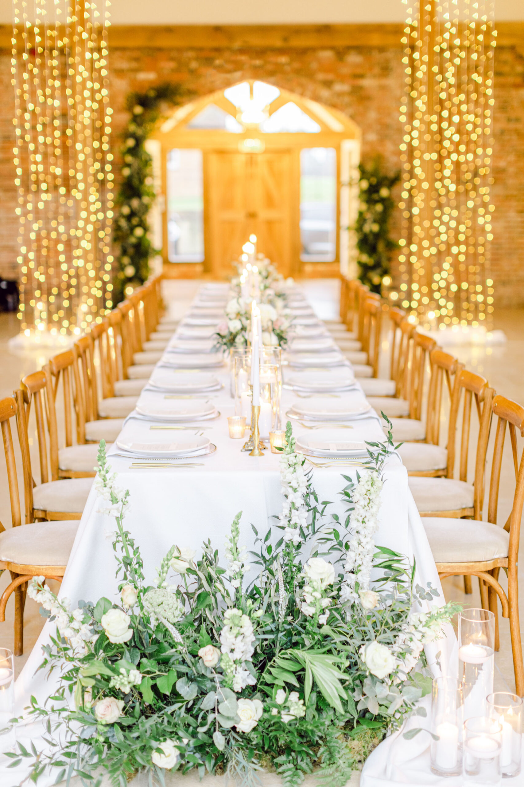 Long banquet style luxury wedding table with lots of greenery and candles down the centre line. The table is highlighted by fairy light candies in the background. Image by Natalie D Photography
