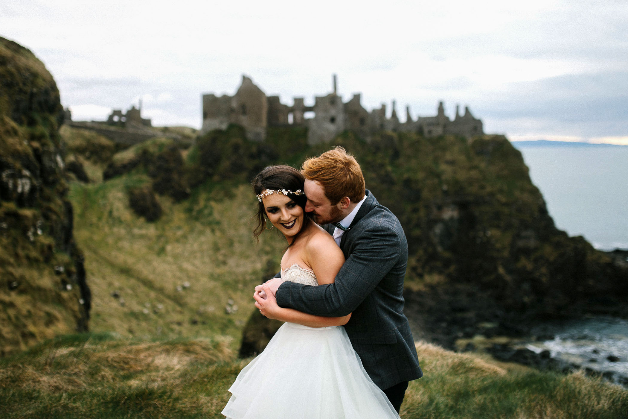 A bride and groom in the countryside near a ruined old building. She's smiling as he hugs her. By Luke Flint Photography