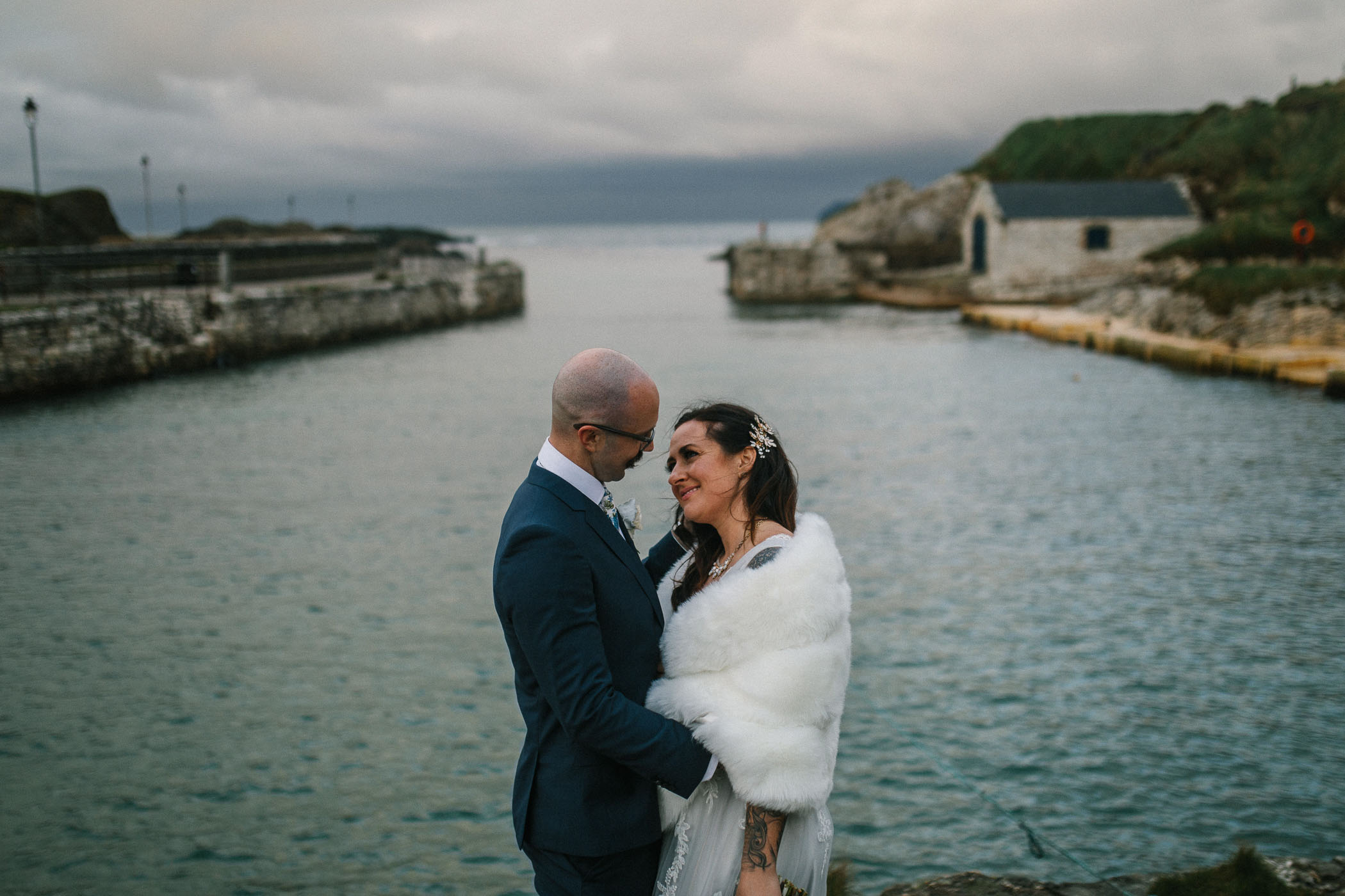 A bride and groom in a harbour with the sea behind them. He's in a blue suit and she's wearing a white faux fur wrap. By Luke Flint Photography