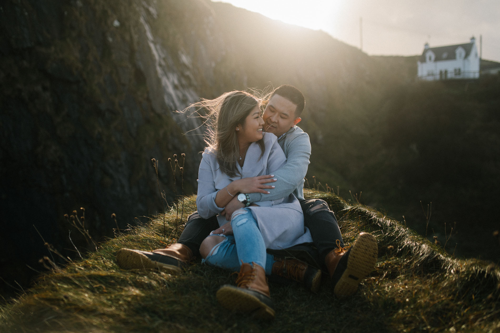 A couple sit snuggled together on a hilltop with a mountain behind them. The sun is flaring in the background. By Luke Flint Photography