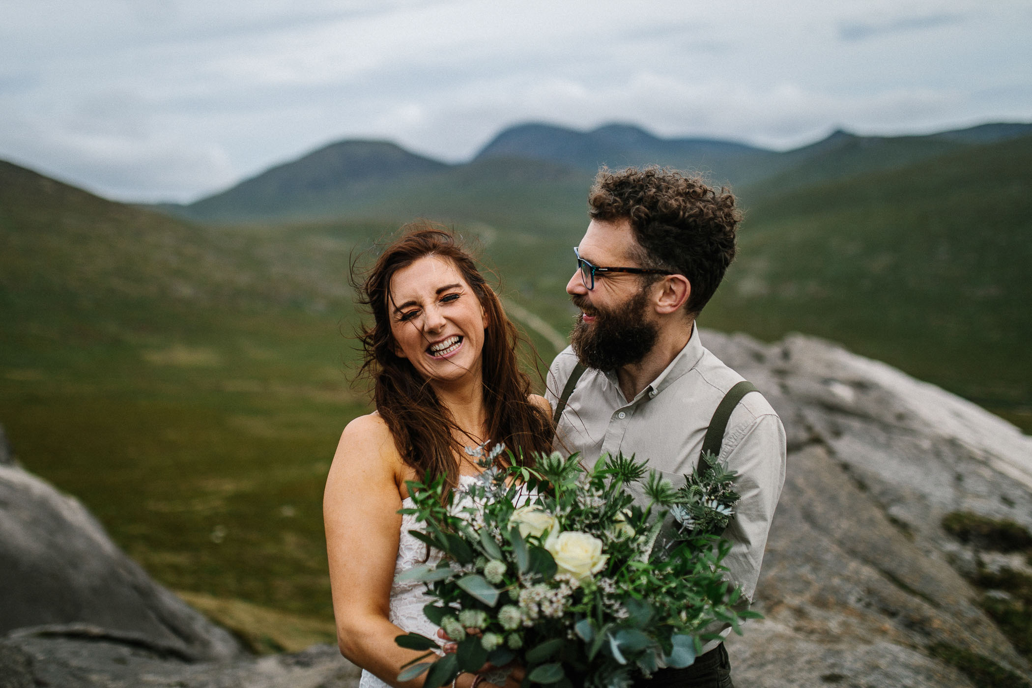 A couple laugh together on their wedding day. They're surrounded by mountains and she's holding a bouquet with lots of greenery. By Luke Flint Photography