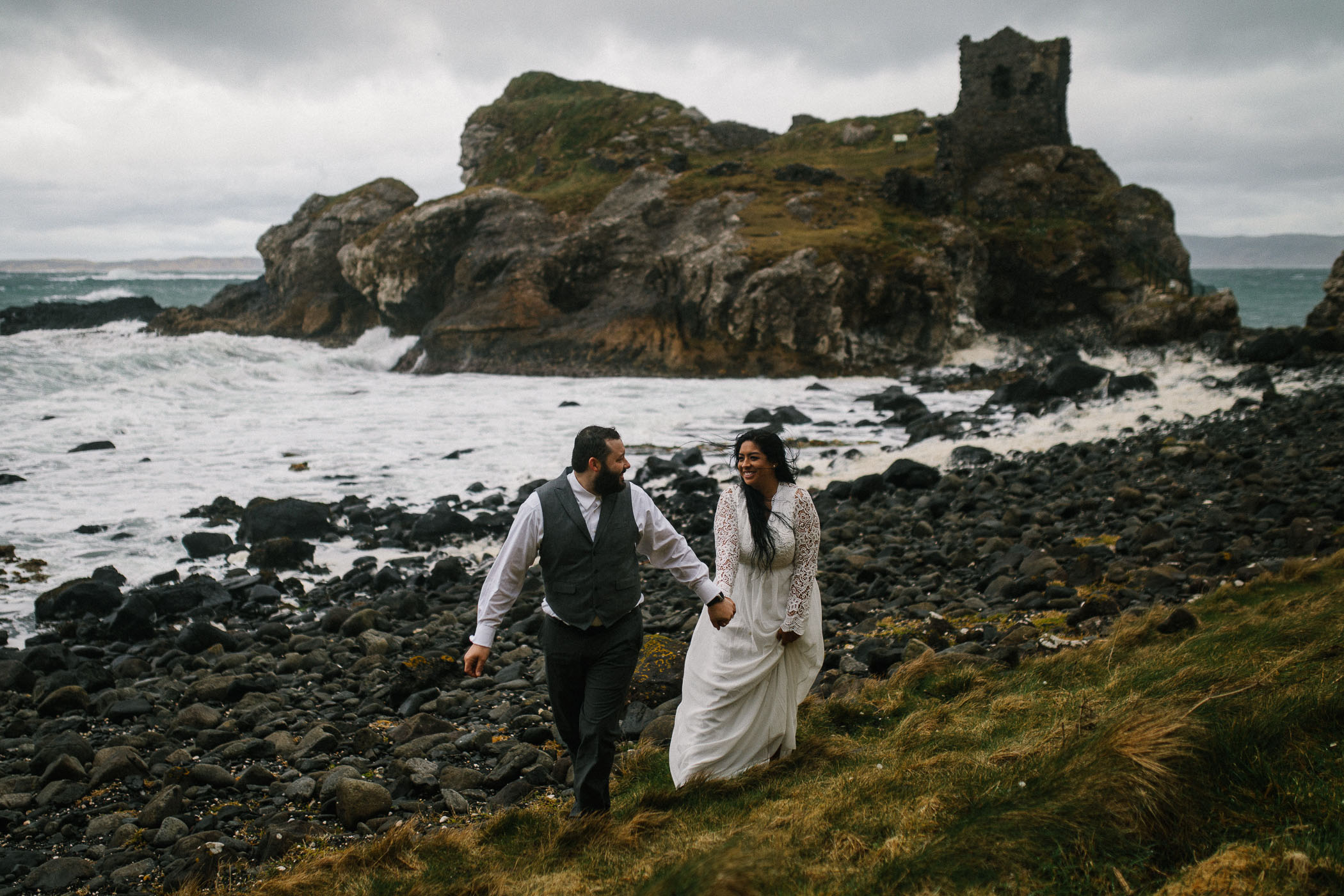 A couple walk along a stony beach with the sea behind them and a ruined building on a rocky outcrop. By Luke Flint Photography