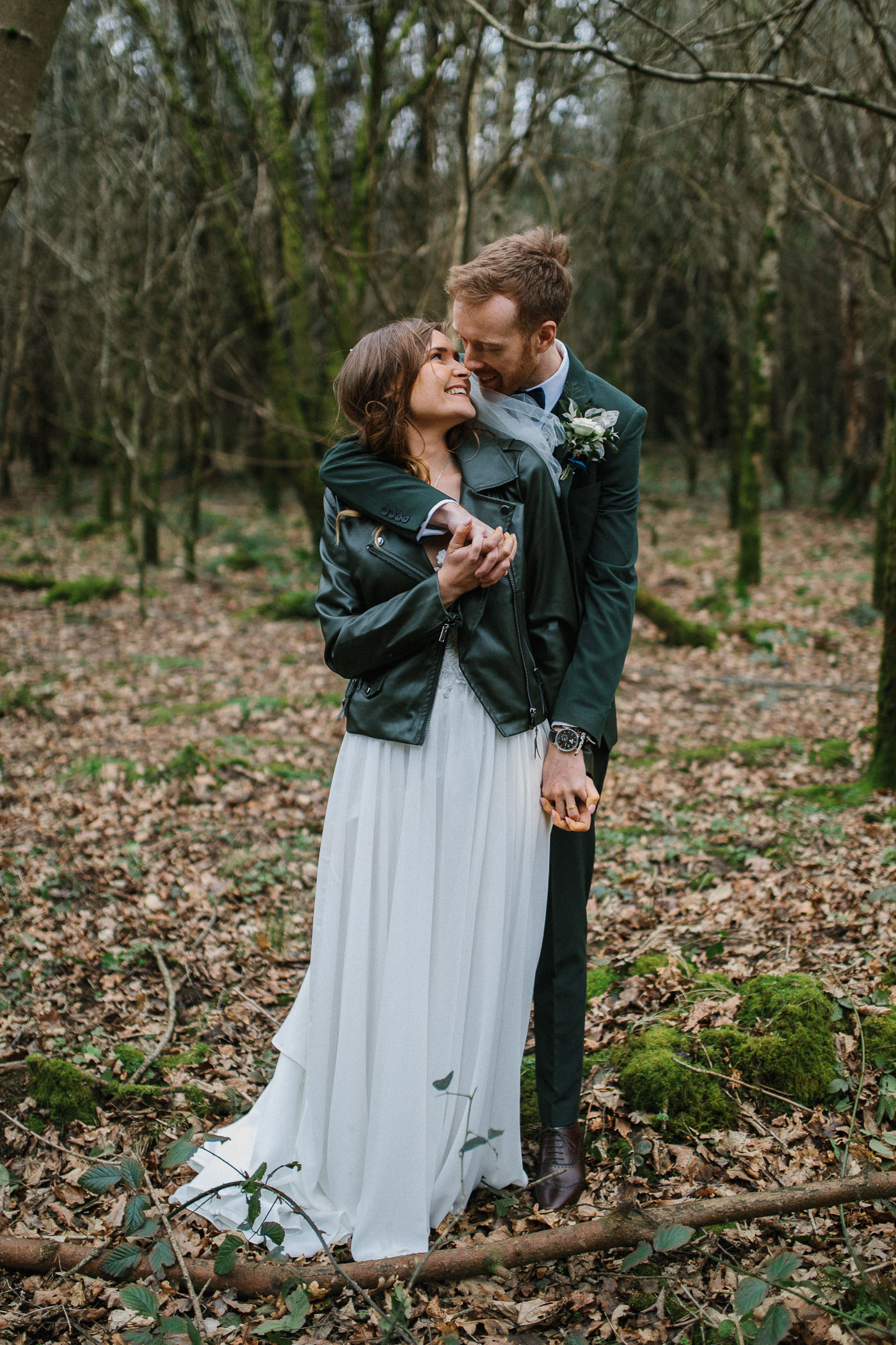 A couple pose for a photo on their wedding day. She's in a long white skirt with a black denim jacket and he's wearing a suit with a button hole. By Luke Flint Photography