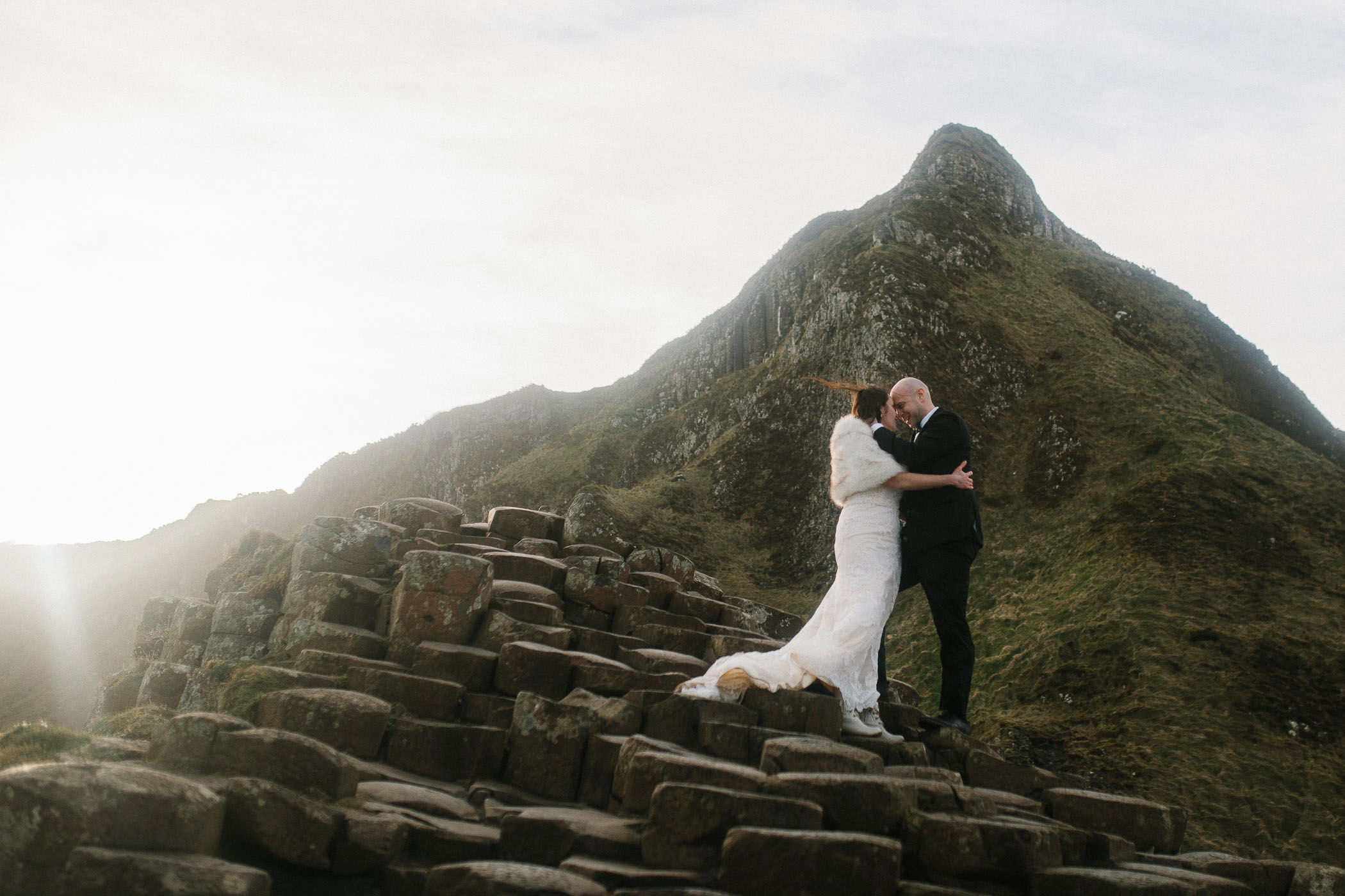 Newlyweds on a rocky outcrop with a mountain in the background and winter sunshine setting behind them. By Luke Flint Photography