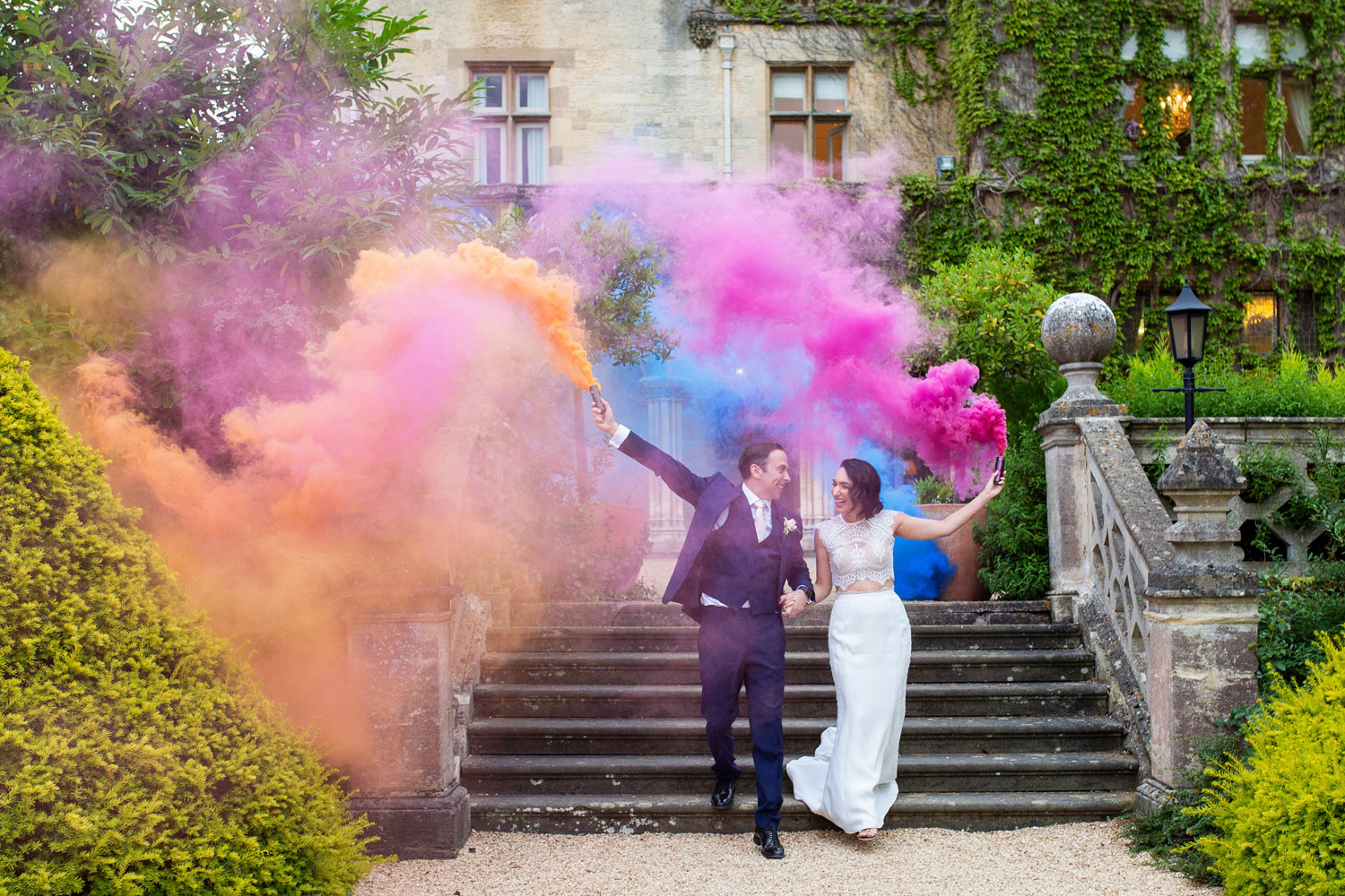 Paar wedelt mit orangefarbenem, blauem und rosafarbenem Rauch.  Er trägt einen Anzug und sie ein trendiges Hochzeitskleid.  Von Hochzeitsfotograf Martin Dabek aus Bristol