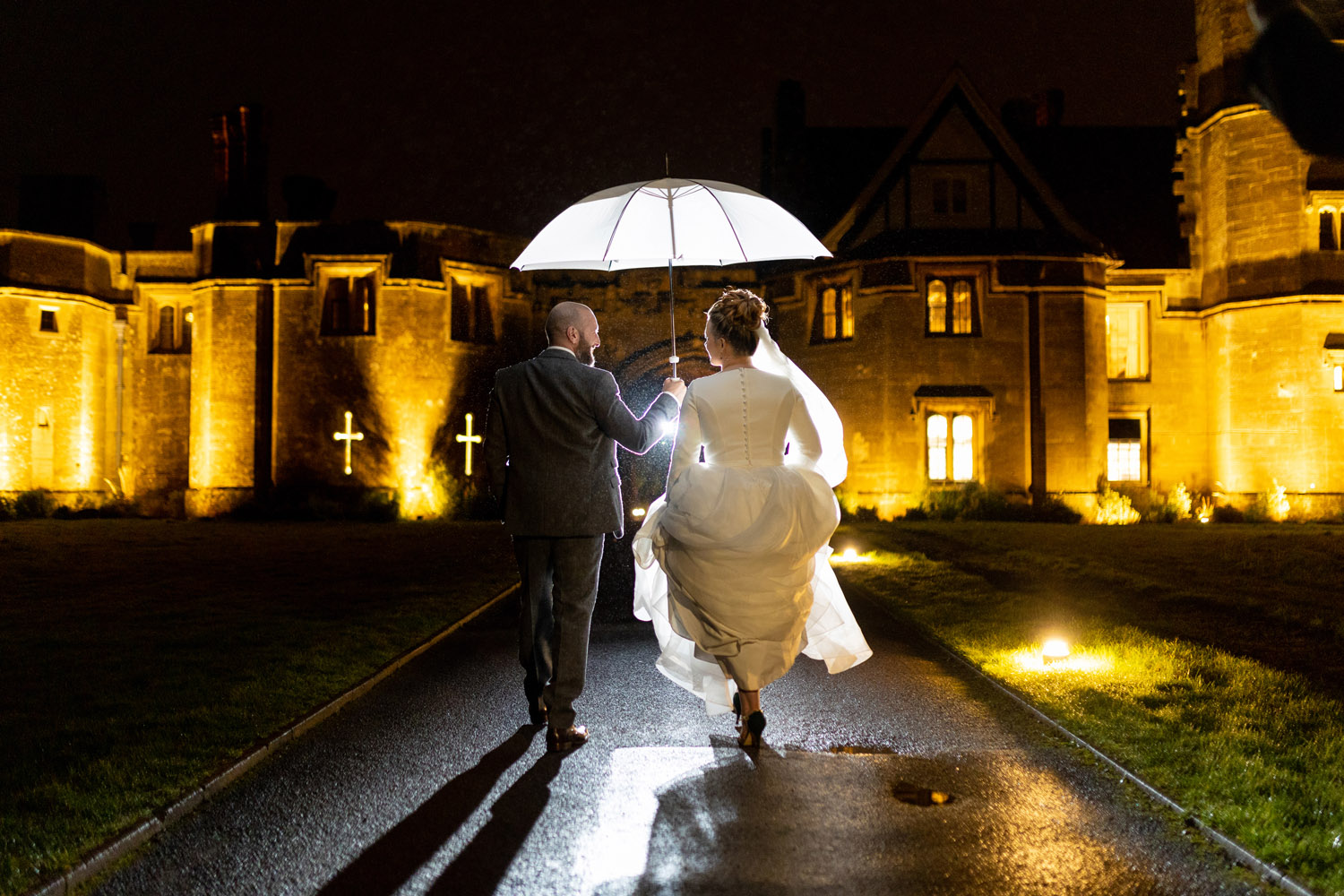 A couple walk down the driveway of a wedding venue in the rain. Their white umbrella is beautifully lit and the house in the background has a golden glow. Martin Dabek Photography