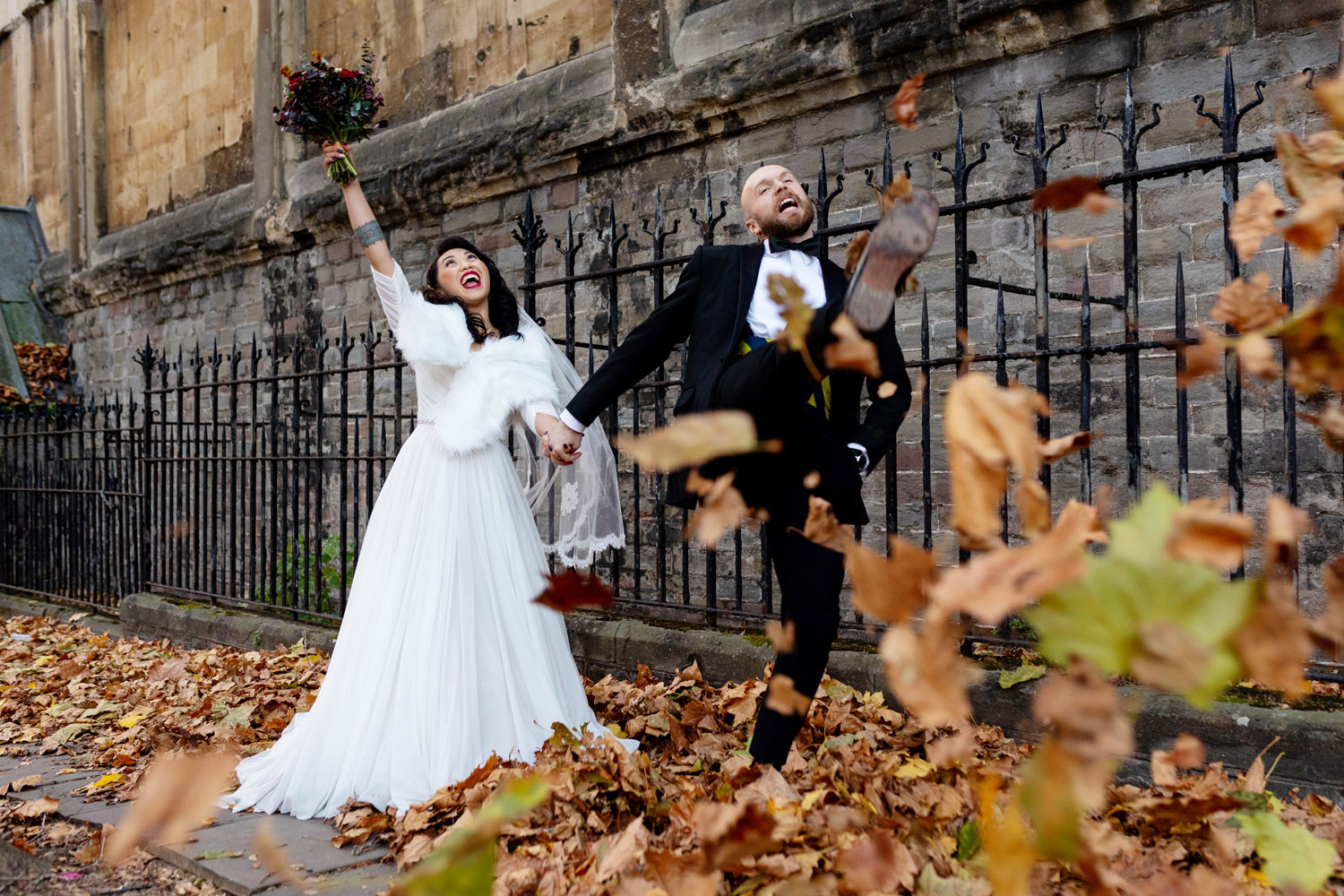 Playful wedding photography of newlyweds kicking autumn leaves. The bride holds a bouquet high in celebration. The groom's just kicked leaves towards the camera. With Martin Dabek Photography