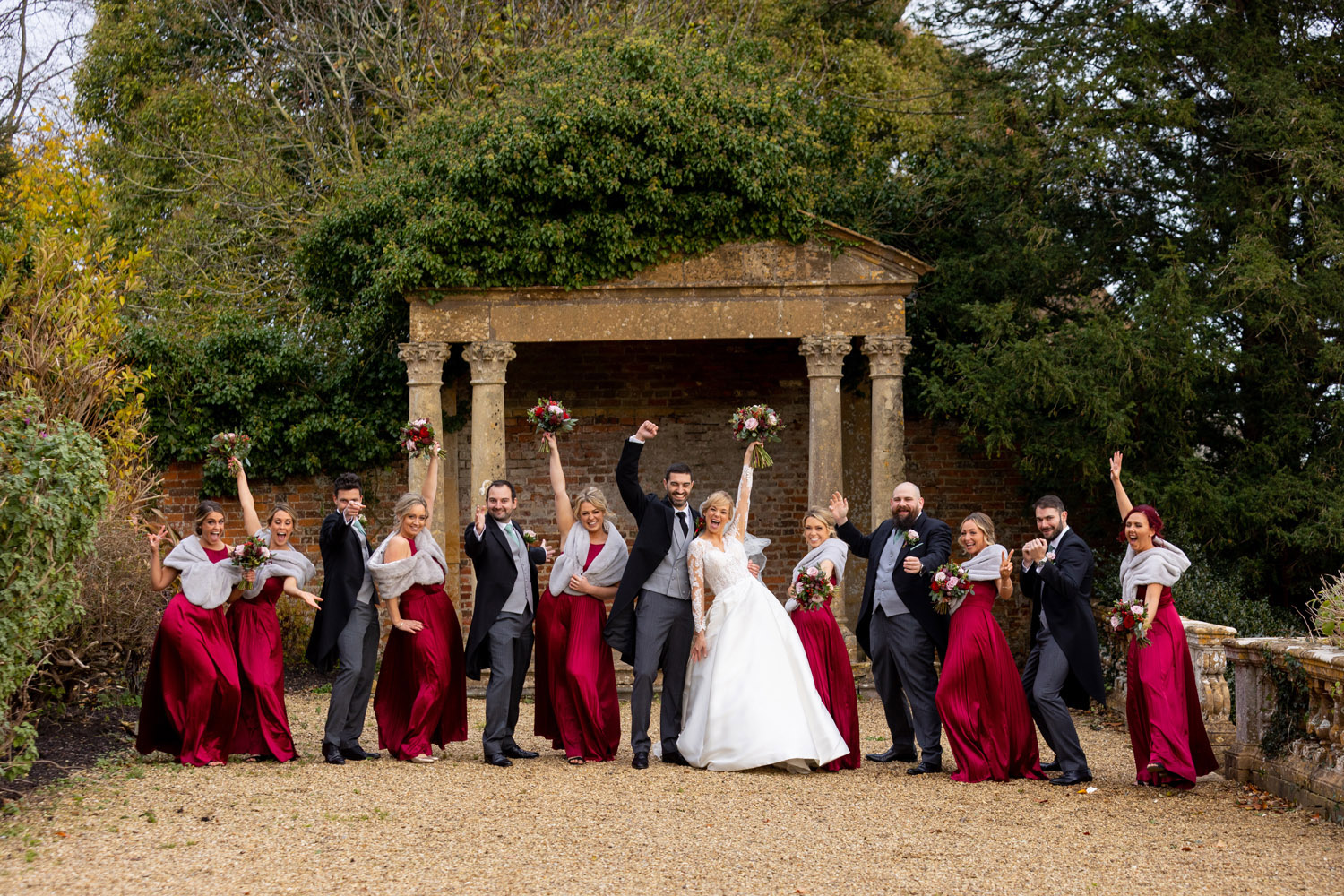 A wedding group shot with 12 people including the bride and groom, bridesmaids in dark red and the bride holding her bouquet high. By Martin Dabek, who's a Bristol wedding photographer