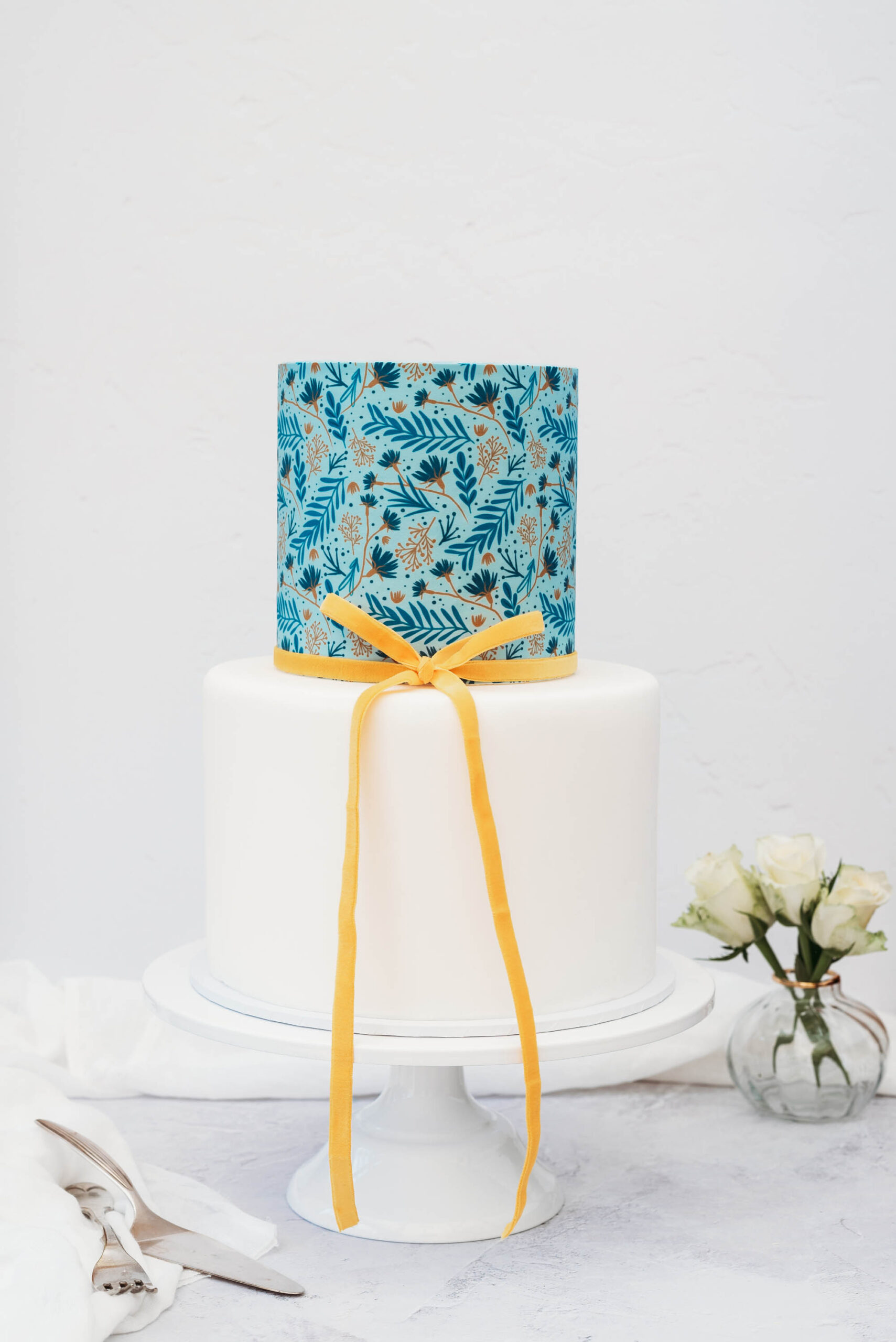 printed botanical wedding cake with two tiers. The top tier has a modern leaf and flower illustration in blues, with a thin gold ribbon around, and the bottom tier is simple white. Cakes by Tasha