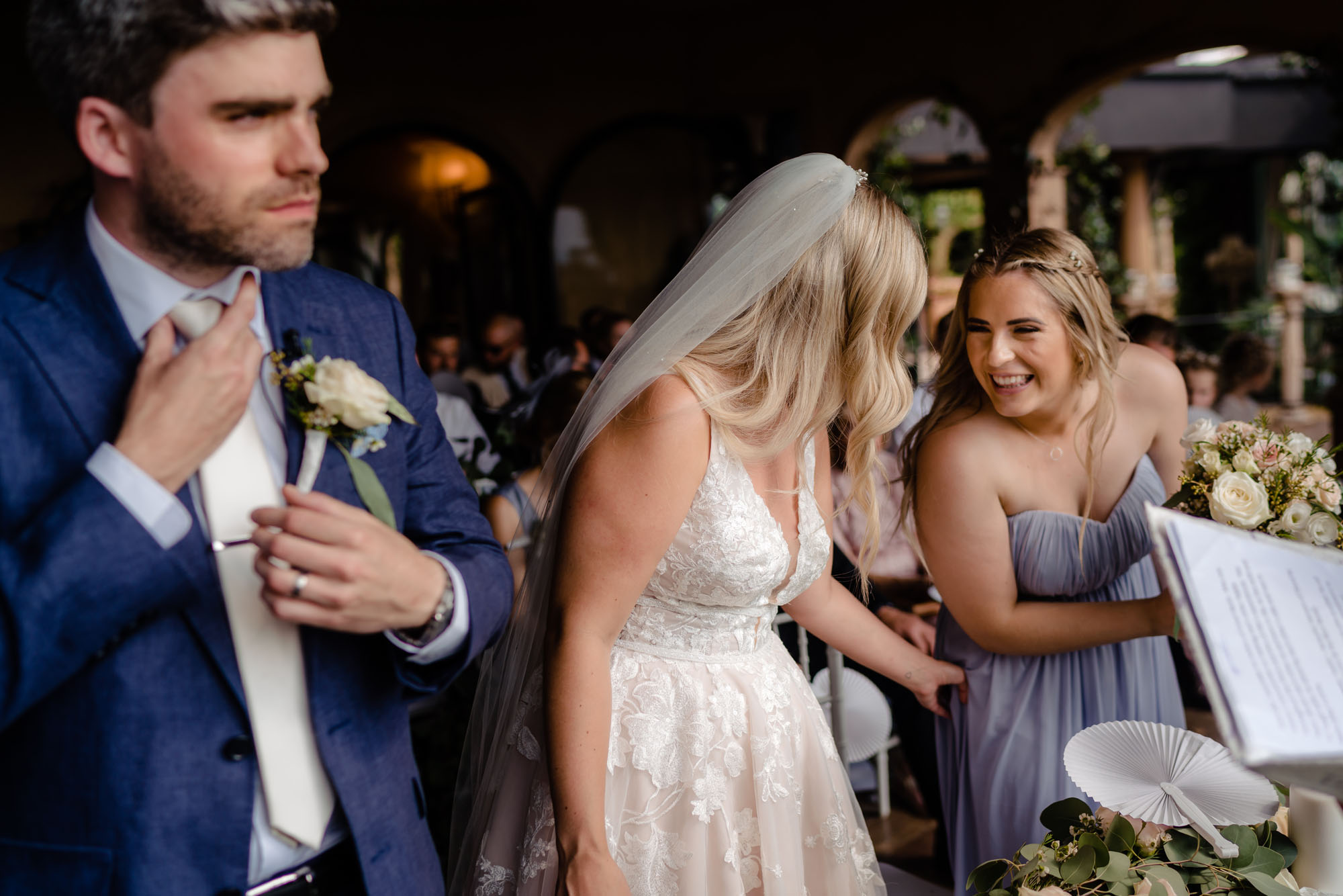A bride chats to her bridesmaid while the groom by her side straightens his tie. By Liam Collard Photography