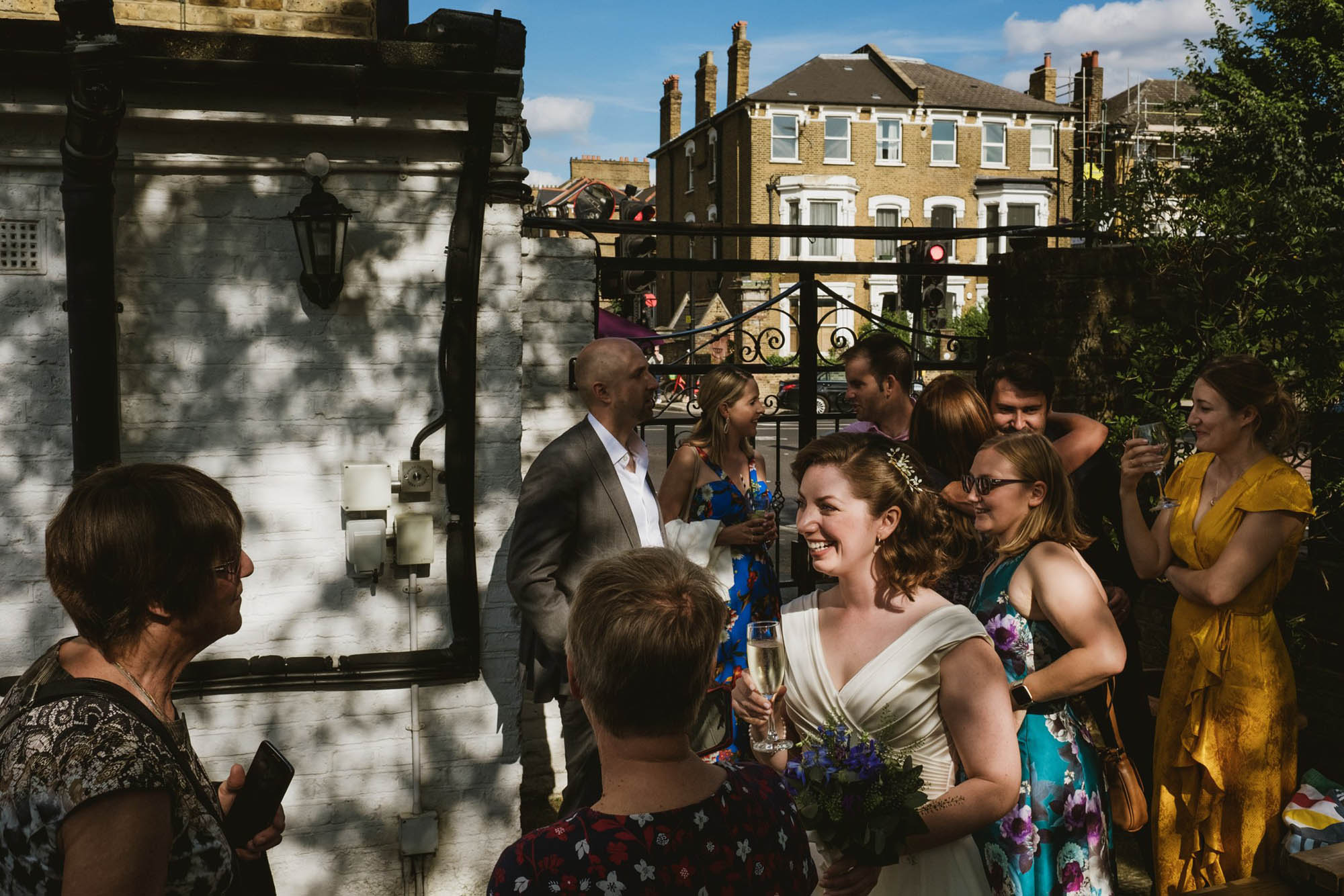 A bride surrounded by friends and family chatting in dappled sunshine at the gates of the wedding venue. By York Place Studios