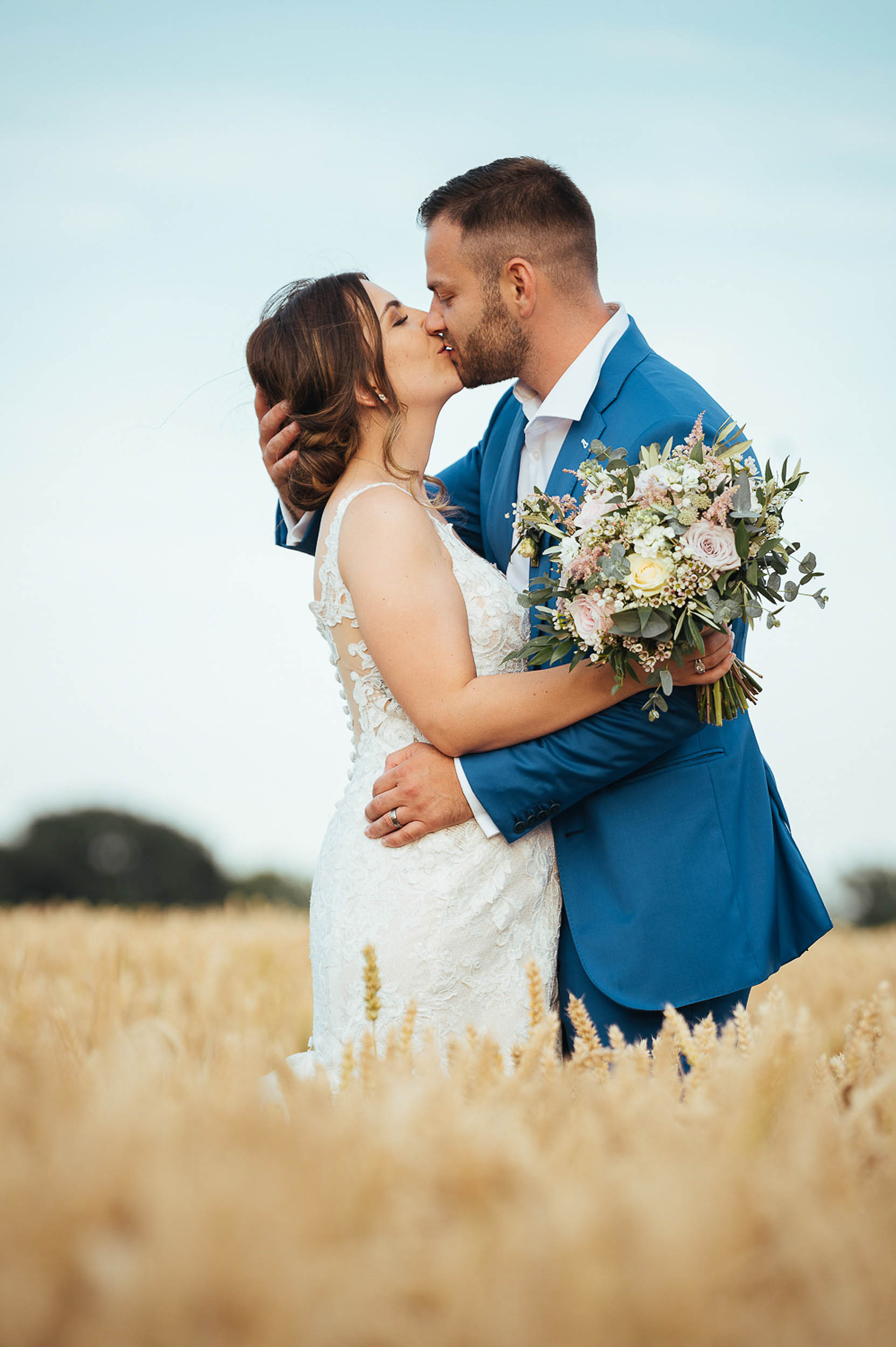 Newlyweds kiss for the camera! He's holding the back of her head with his arm around her waist, and she's carrying a bunch of white boho flowers - Ben Pipe Photography