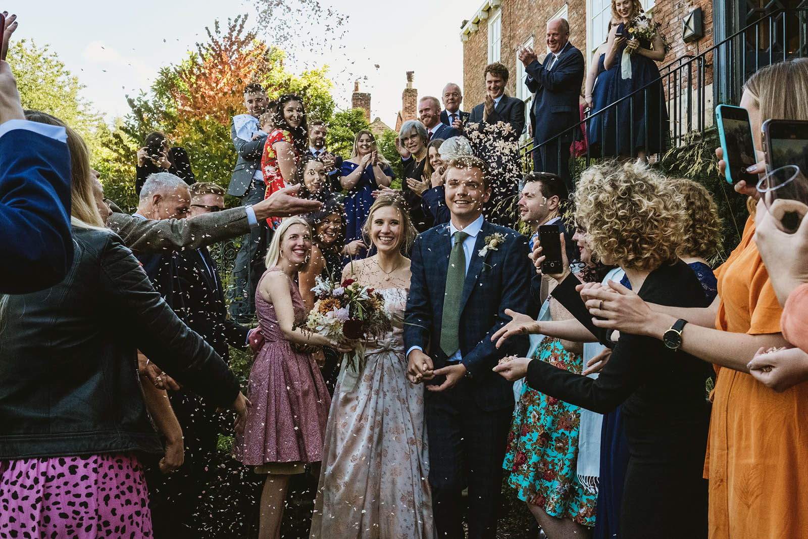 Newlyweds leaving the venue in a cloud of dried petal confetti. Unposed and natural wedding photography with real smiles, by York Place Studios