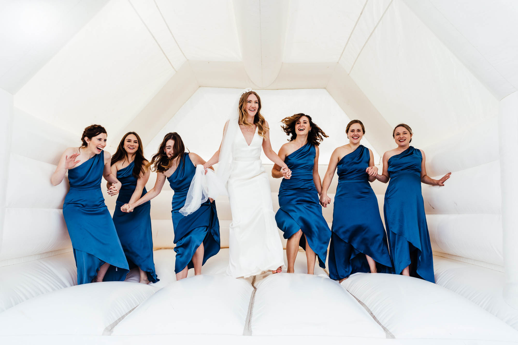 Bride with her bridesmaids laughing as they play in a white bouncy castle - by Hannah Hall Photography