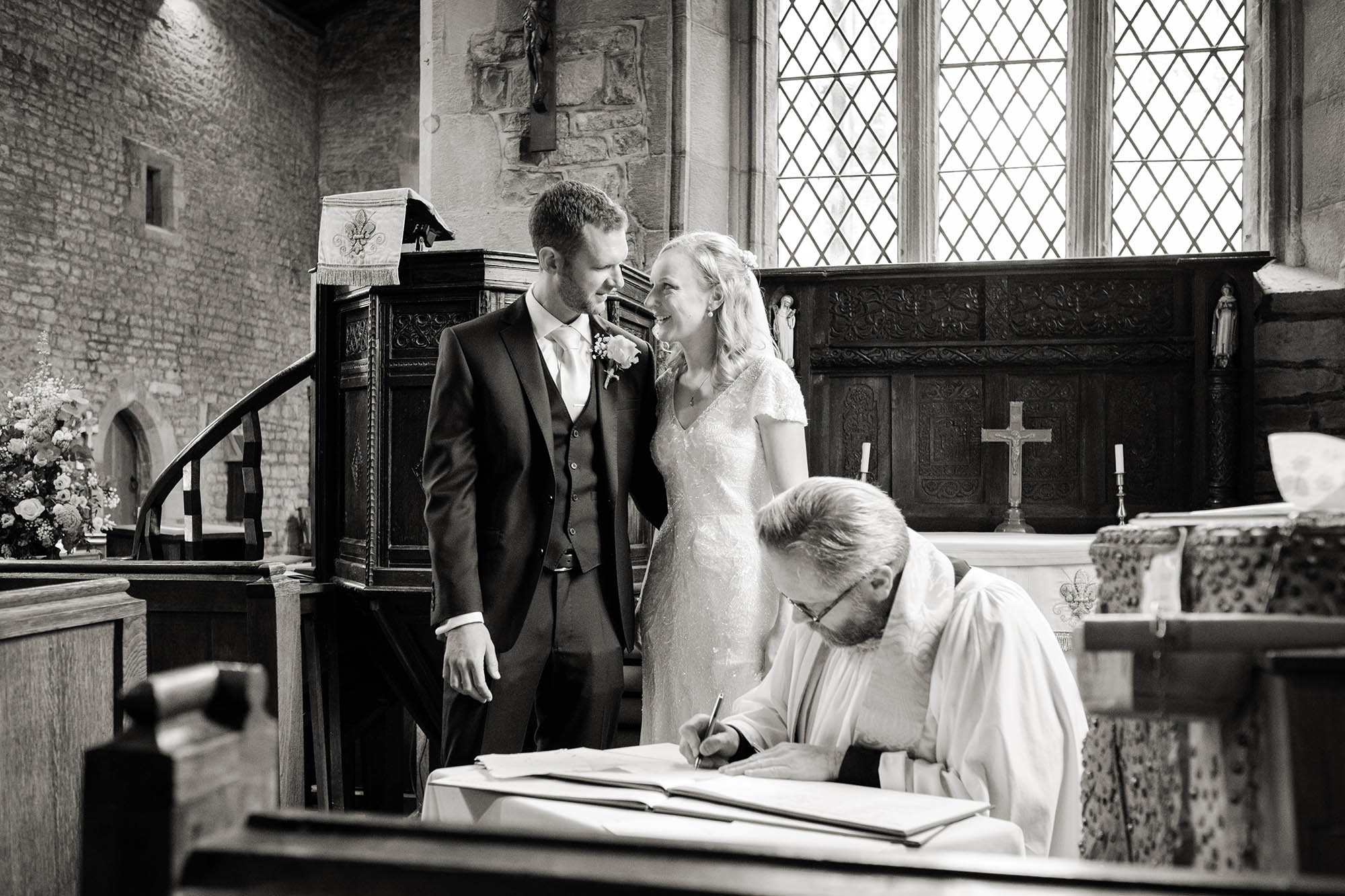 Newlyweds share a moment with a loving glance as the priest signs the marriage register - Sheffield wedding photographer John Mottershaw